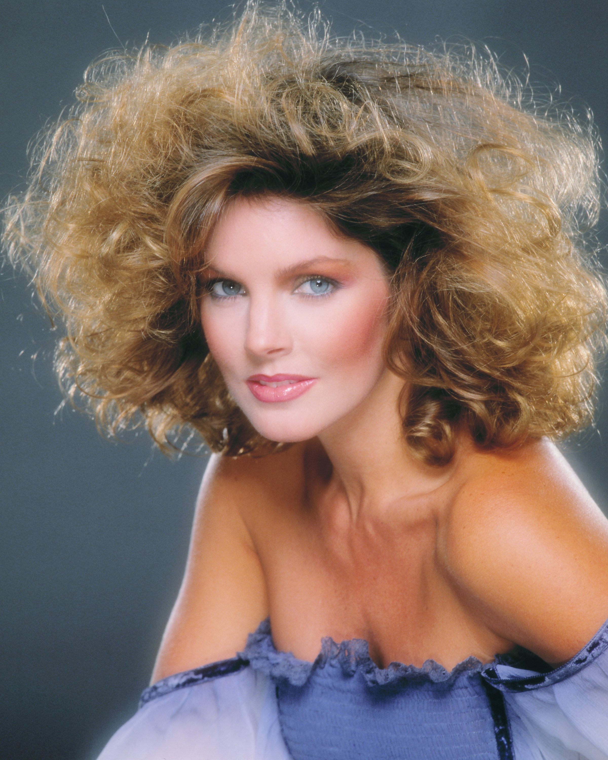 Priscilla Presley poses for a studio portrait in Los Angeles, California, in 1979, wearing an off-the-shoulder periwinkle-blue top with delicate ruffled lace trim, her highlighted hair styled in voluminous feathered curls. | Source: Getty Images