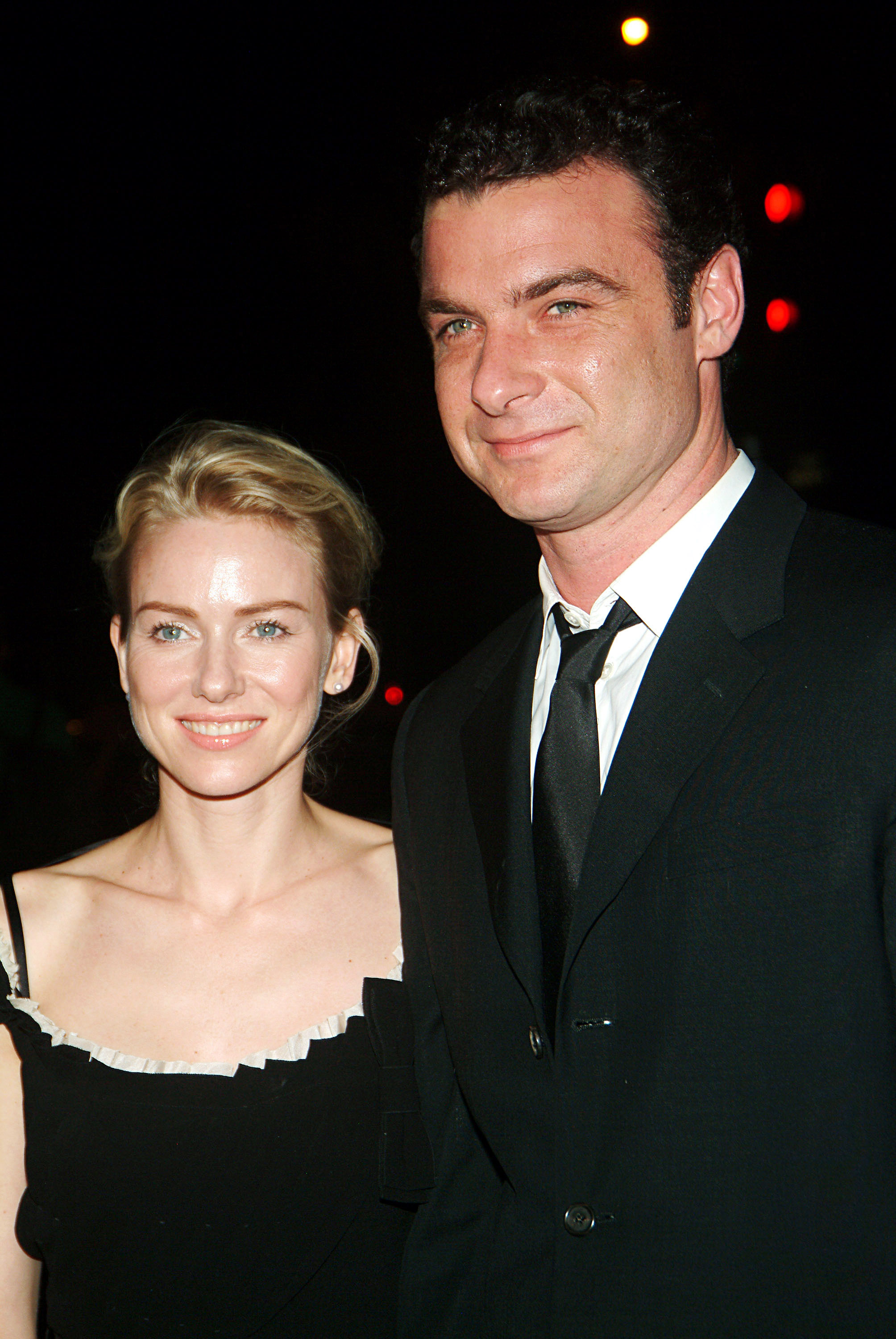 Naomi Watts smiles softly toward the camera while Liev Schreiber stands beside her with a relaxed confidence, the pair illuminated against a dark backdrop dotted with glowing city lights.