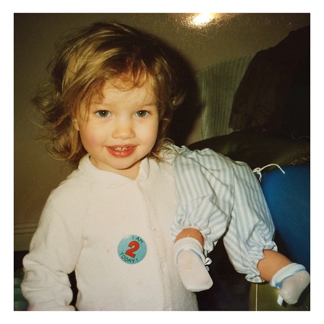Rosie Alice Huntington-Whiteley smiles in a childhood snapshot, wearing a white outfit with a “I am 2 today” badge and holding a doll. | Source: Instagram/rosiehw