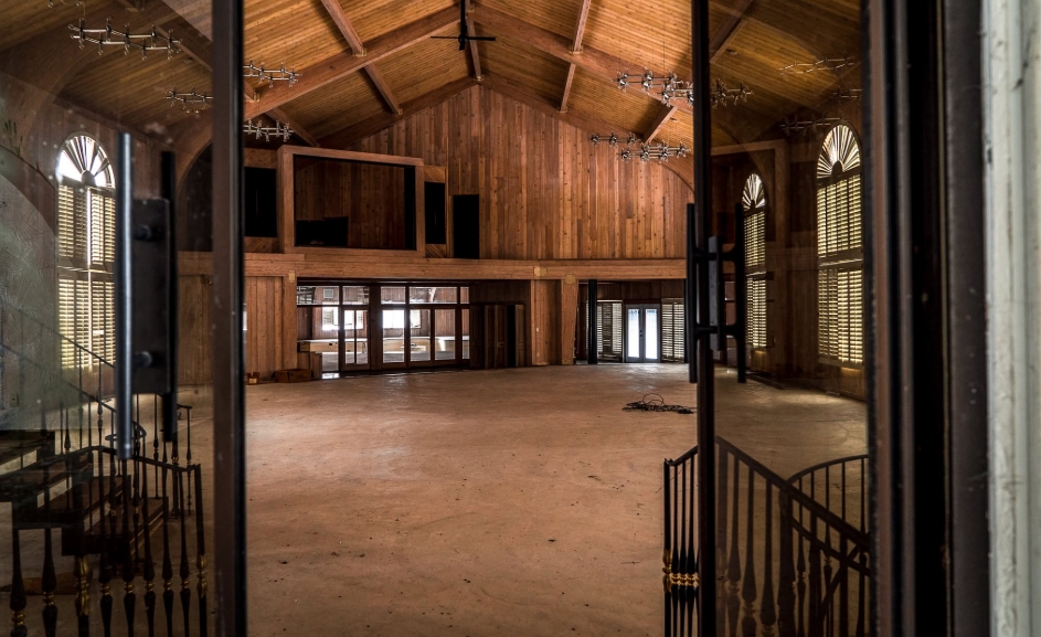 Wood paneling stretches from floor to ceiling inside one of the largest spaces at Mike Tyson's onetime Ohio mansion, with tall shuttered windows and a vaulted roof emphasizing its considerable height. Multiple lighting fixtures remain suspended overhead, while an upper section overlooks the open floor below, illustrating the grand scale that characterized the boxer's sprawling residence during his ownership. | Source: Facebook/Johnny Joo