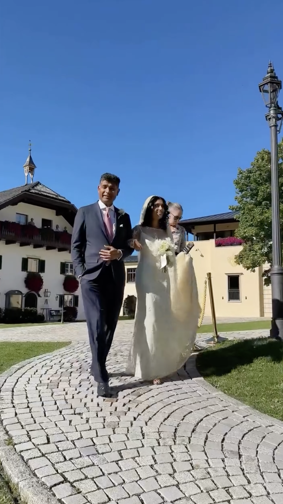 Sheela Newley walks with a loved ones across the stone path in her lace wedding gown and veil. She holds a classic bouquet, her expression joyful as she approaches the ceremony location. | Source: Instagram/sheelanewley