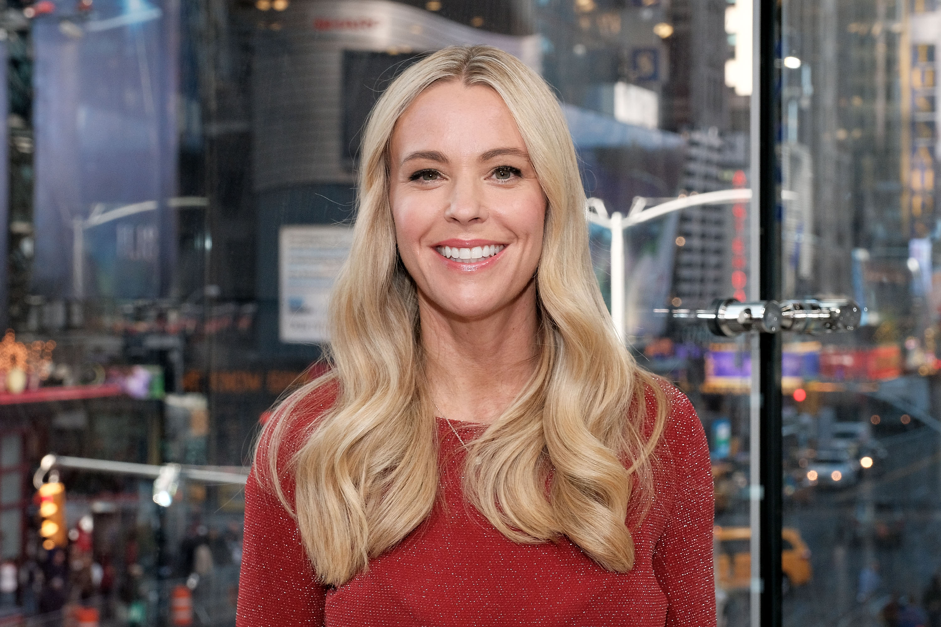 Visiting "Extra" in Times Square, Kate Gosselin smiles in a red top. Her long blonde waves and natural makeup highlight a relaxed, polished look as she appears upbeat against the backdrop of the bustling city lights.