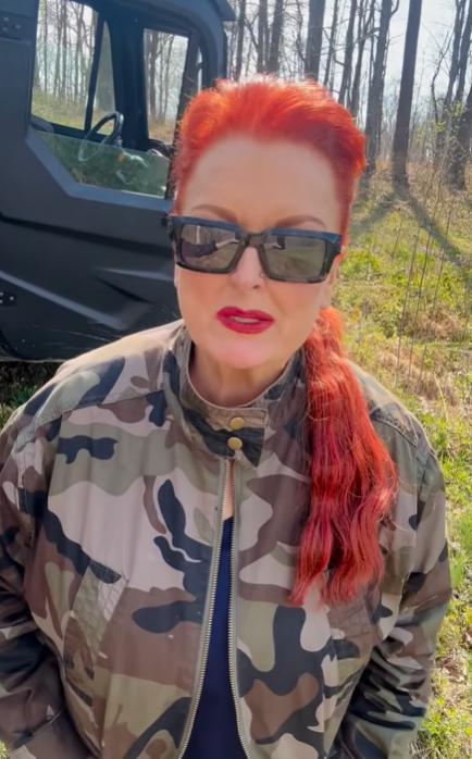 Wynonna appears with her bright red hair pulled back, wearing a camouflage jacket beside an off-road vehicle. Her outdoor clothing offers a contrast to the rhinestones and stage outfits associated with her music career. | Source: Youtube/Wynonna