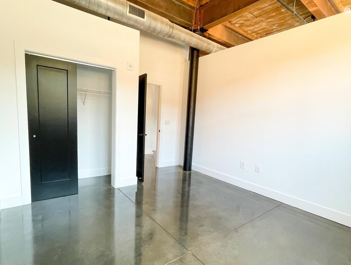 An unfurnished loft reveals the property's industrial bones, from the polished concrete floor and exposed wood ceiling to the prominent ventilation duct and structural column. | Source: Facebook/Judson Mills Loft