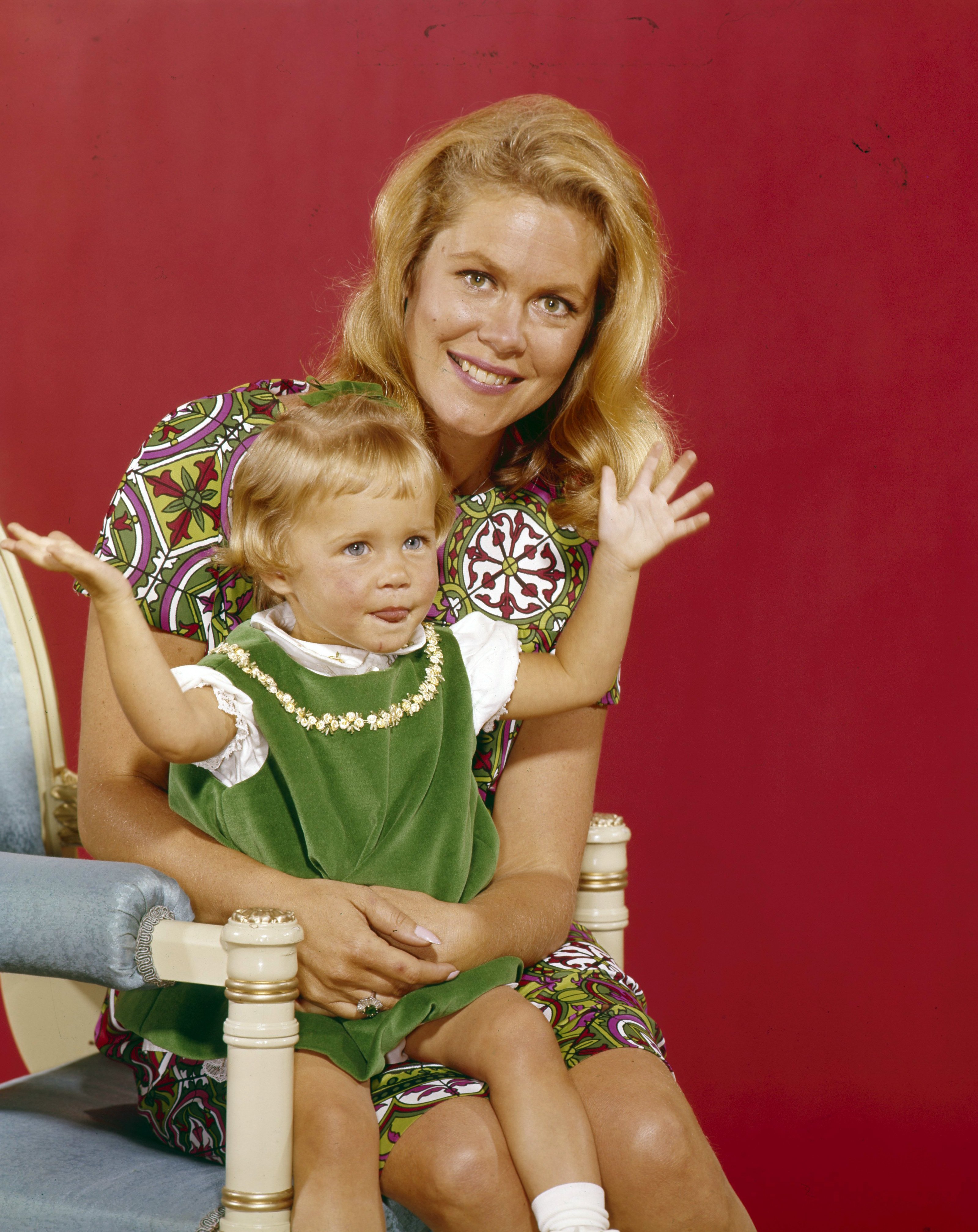 A very young Erin Murphy sits with Elizabeth Montgomery in a "Bewitched" cast portrait, with Murphy raising both hands as Montgomery smiles behind her. The image captures Murphy near the beginning of the role that would define her childhood career and offers an early point of comparison with her 2026 appearance.