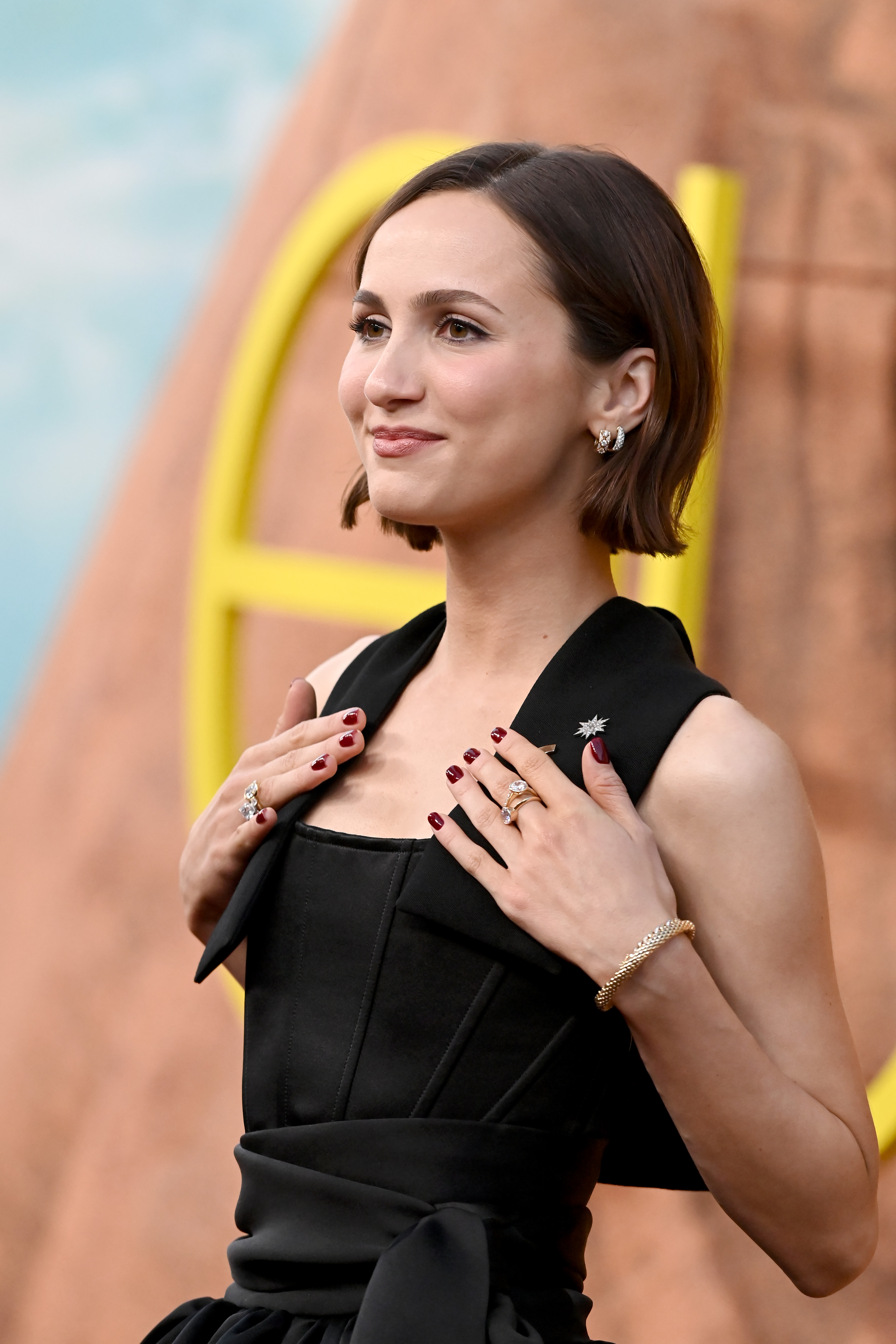 At the premiere, Maude Apatow keeps it polished in a structured black look, featuring a corset-style bodice and sharp, tailored lapels. Her sleek bob and soft glam makeup add a modern, understated elegance. Delicate rings, a gold bracelet, and deep red nails bring a subtle yet striking finish.