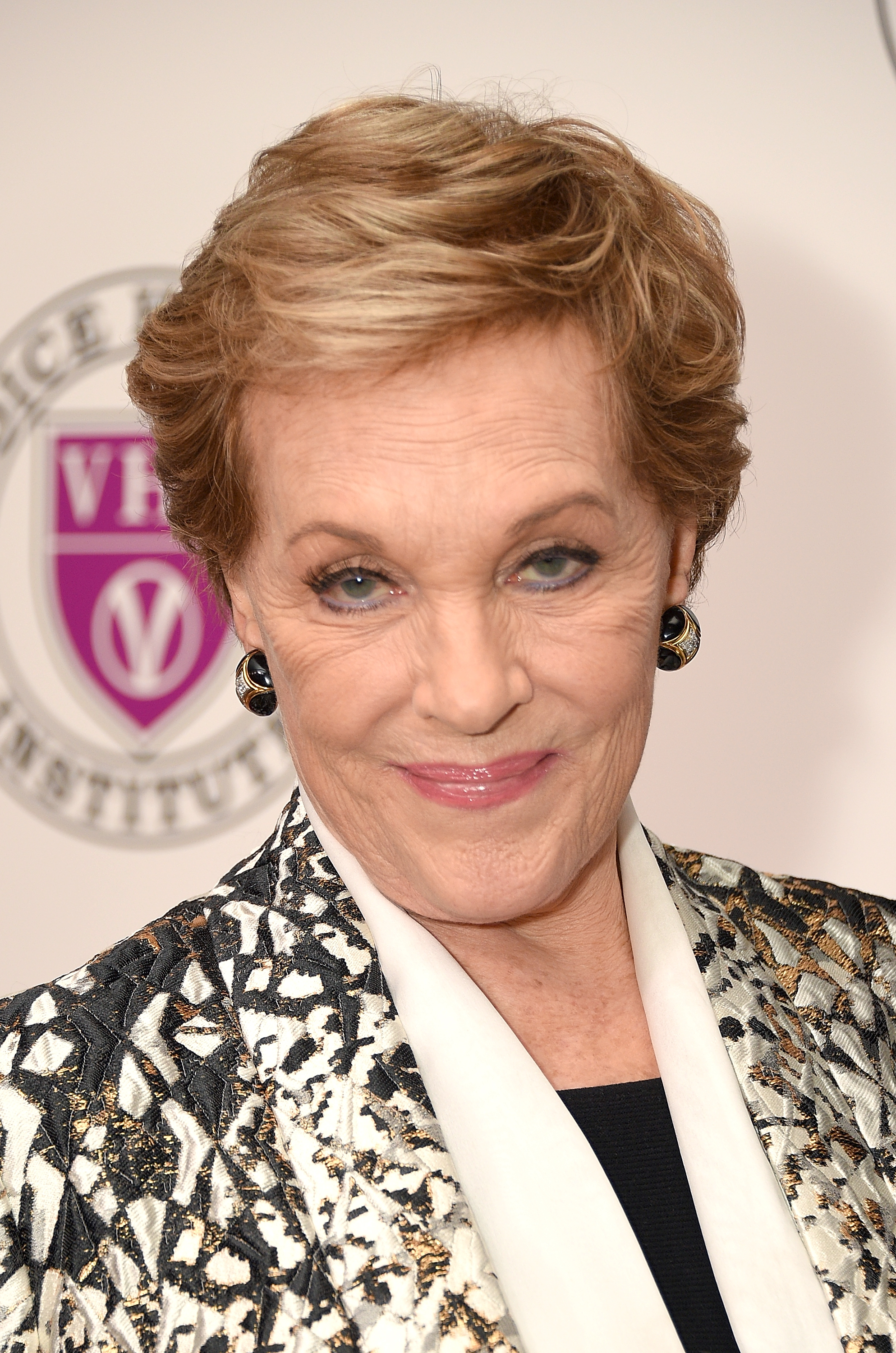Julie Andrews smiles warmly on the red carpet at the "Raise Your Voice" concert honoring her at Alice Tully Hall, Lincoln Center, on March 5, 2018, in New York City. The screen and stage legend looks radiant in an intricately textured black, gold, and white patterned jacket layered over an ivory shawl collar and dark blouse, complemented by elegant black-and-gold enamel stud earrings and a soft pink lip.