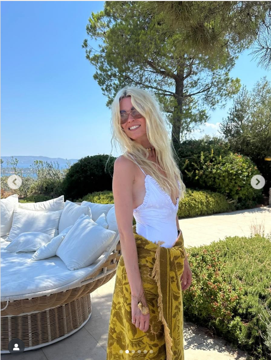 Claudia Schiffer beams with elegance on her birthday, posing in a chic white top and golden patterned wrap against a serene outdoor backdrop. With sunlit hair and a joyful smile, she celebrates timeless beauty and grace | Source: Instagram/claudiaschiffer