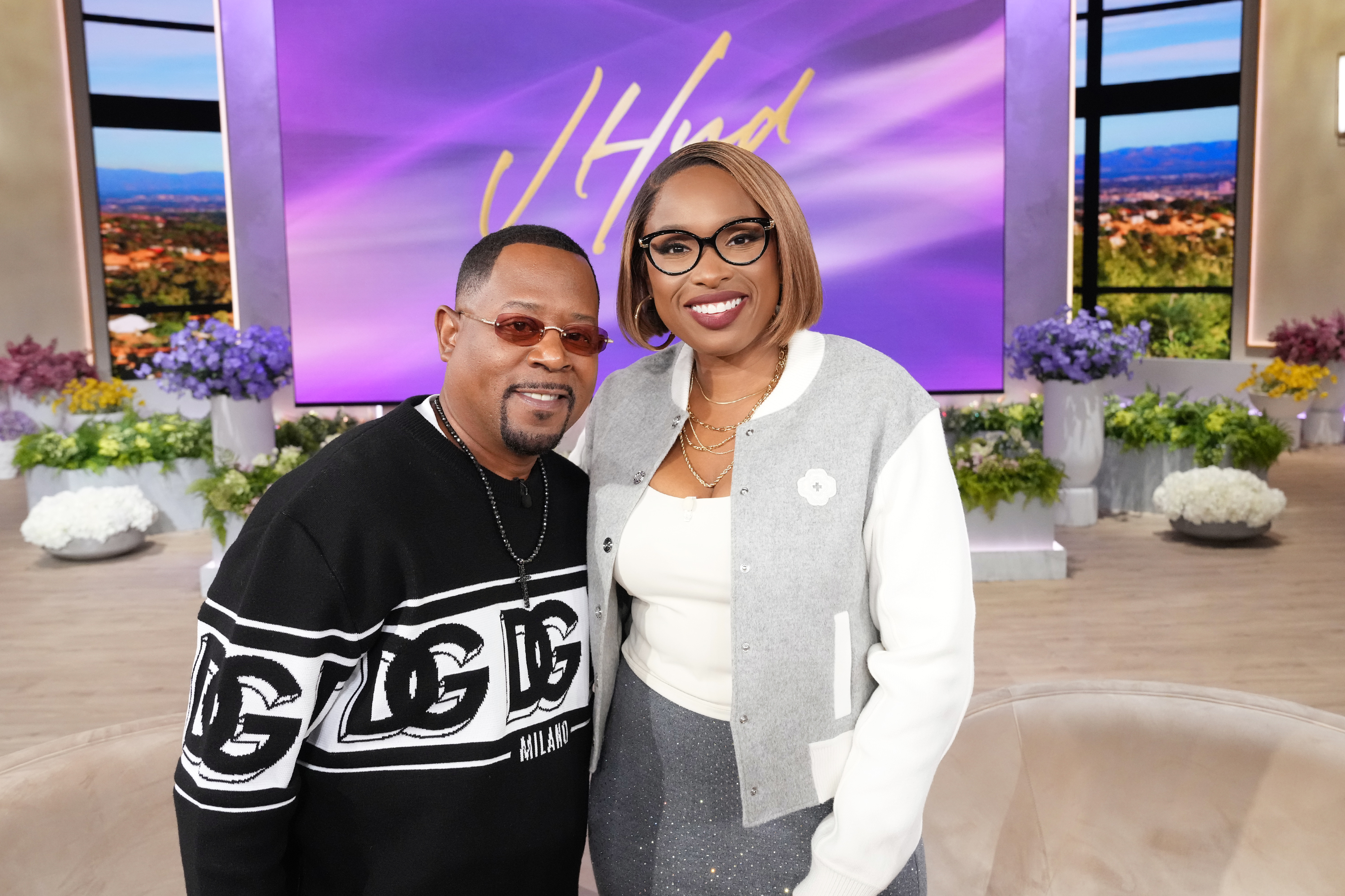 Appearing on "The Jennifer Hudson Show," Martin Lawrence stands next to Jennifer Hudson in a brightly lit studio set. He smiles warmly, dressed casually in a patterned sweater, while she stands close beside him in a chic outfit.