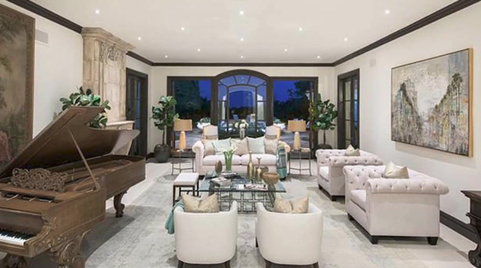 The elegant formal living room and music salon features an ornately carved upright grand piano, plush tufted Chesterfield-style armchairs, and a carved limestone fireplace, accented by dark crown molding and grand arched French doors leading to the rear patio. | Source: YouTube/Taste of Country