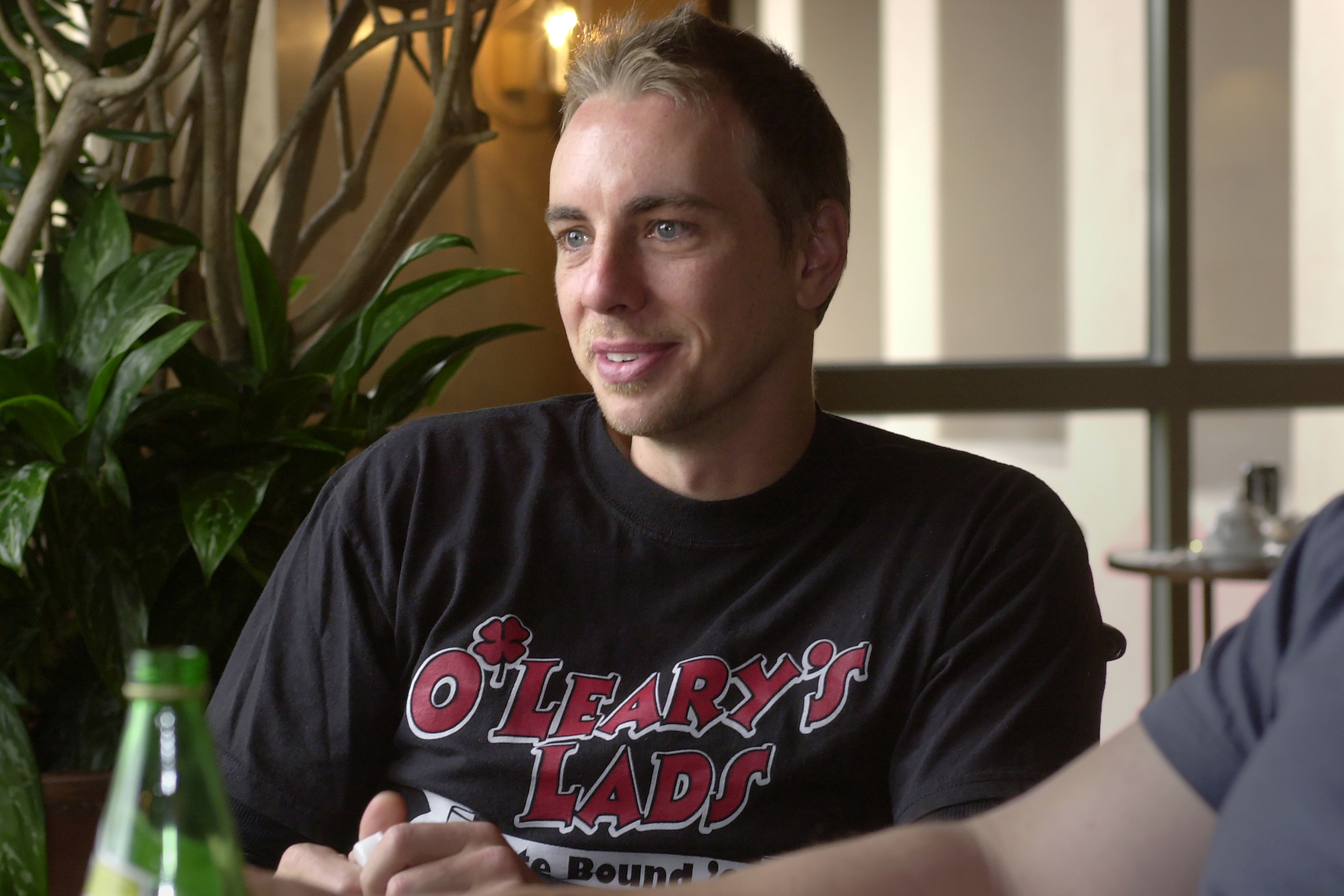 Dax Shepard chats during an interview, wearing a bold graphic T-shirt and an easy smile. Seated near a sunlit window, he brings a casual charm and natural charisma to the conversation.