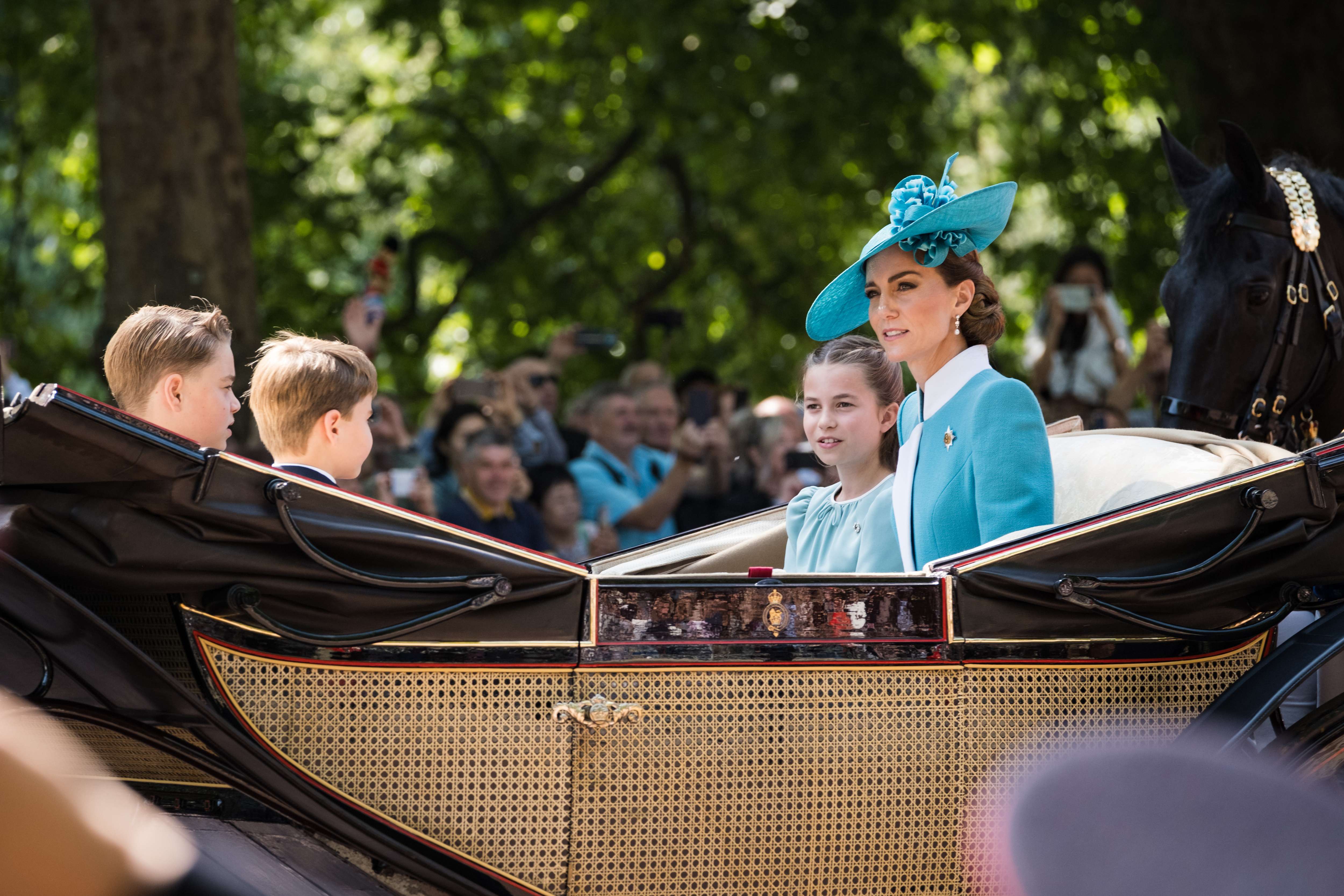 Princess Charlotte, Princess Catherine, Prince George, and Prince Louis ride in the royal carriage as people look-on and take photos of the mother with her children.