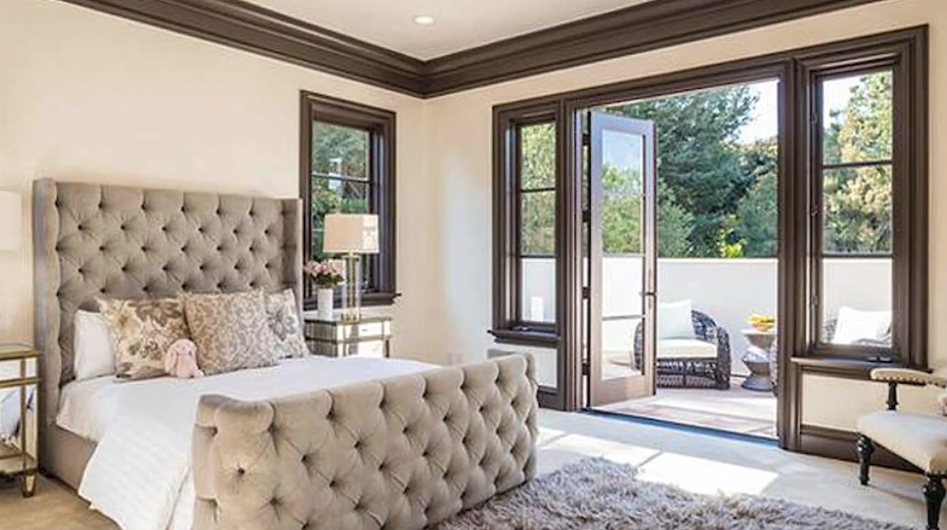 A spacious guest bedroom pairs soft beige walls and rich espresso trim with a dramatic floor-to-ceiling tufted bed frame, opening through glass French doors onto a sunny private terrace that overlooks mature trees. | Source: YouTube/Taste of Country