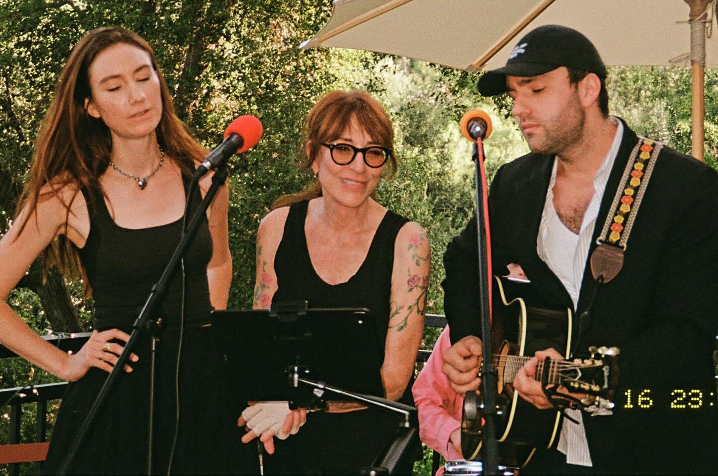 Katey Sagal performed an intimate outdoor musical set alongside her children, Sarah Grace White and Jackson James White, in a photograph shared to social media on September 19, 2024. Flanked by vocal microphones and an acoustic guitar beneath a patio umbrella, the singer-actress smiled warmly in round black glasses and a sleeveless black top displaying her floral arm tattoos, harmonizing with Sarah Grace while Jackson James accompanied them on guitar in a black blazer and baseball cap. | Source: Facebook/Katey Sagal
