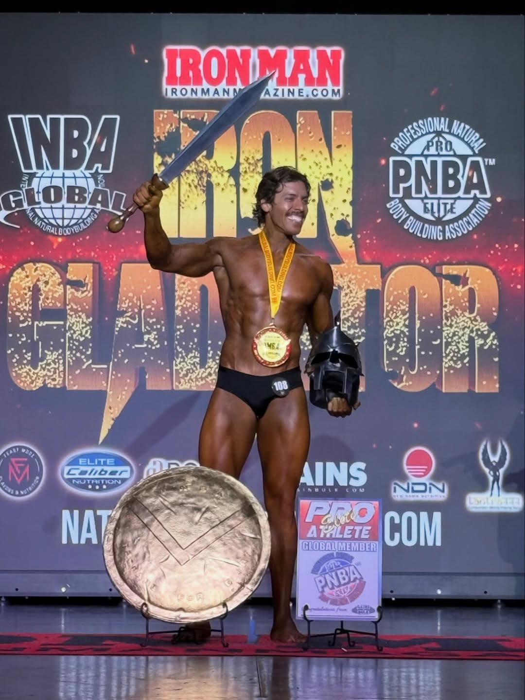 Joseph Baena stands center stage, smiling as he raises a sword overhead while wearing a gold medal and holding a gladiator helmet at his side | Source: Instagram/joebaena