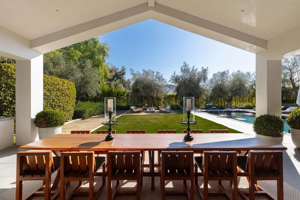 The outdoor dining area provides seating for a large group while remaining sheltered beneath the roofline. From the table, the view extends across the lawn toward the pool and mature landscaping beyond. | Source: Instagram/theshevins