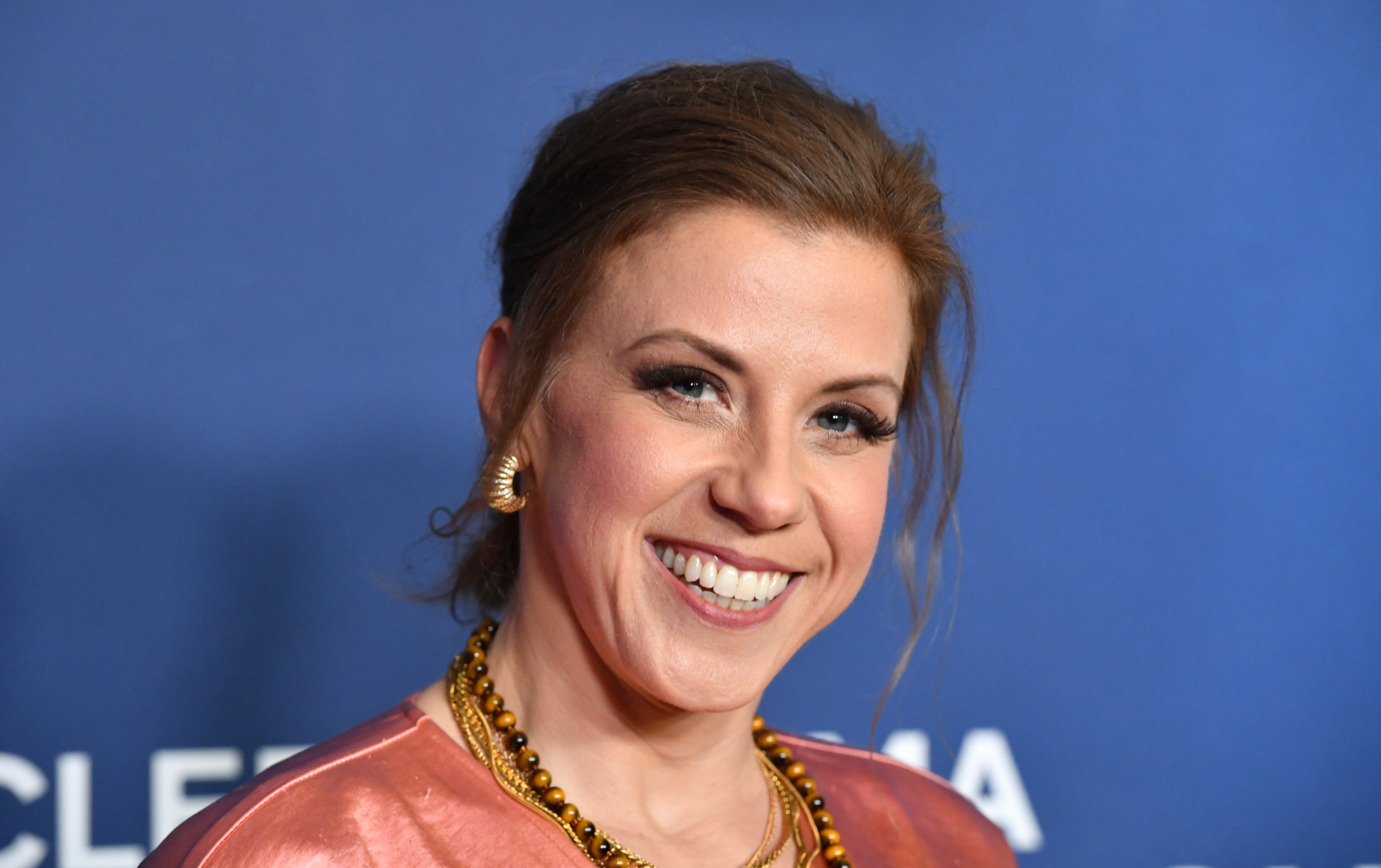 Jodie Sweetin beams on the red carpet at the Scleroderma Research Foundation's "Cool Comedy - Hot Cuisine" fundraiser at the Beverly Wilshire on October 13, 2026. Stepping out in an elegant rose-gold satin gown and styled updo, the actress showcases stunning contemporary glamour decades after charming audiences as little Stephanie Tanner, while continuing an active career across television mysteries and holiday films.