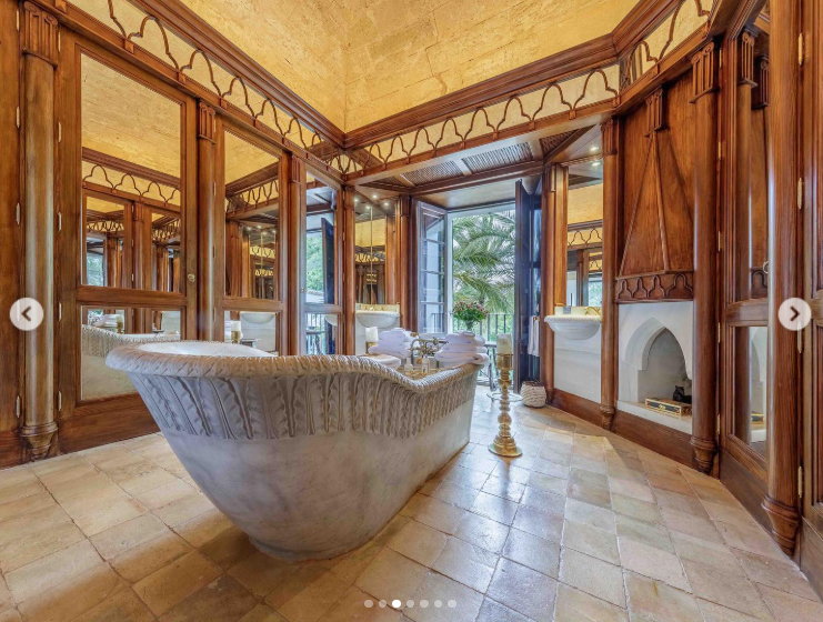 Rich woodwork and mirrored panels surround an ornate tub in one of S'Estaca's bathrooms, while open doors bring in natural light and views of the greenery outside. The intricate interior shows the older decorative character that remains within the estate despite later modernization. | Source: Instagram/robbreport
