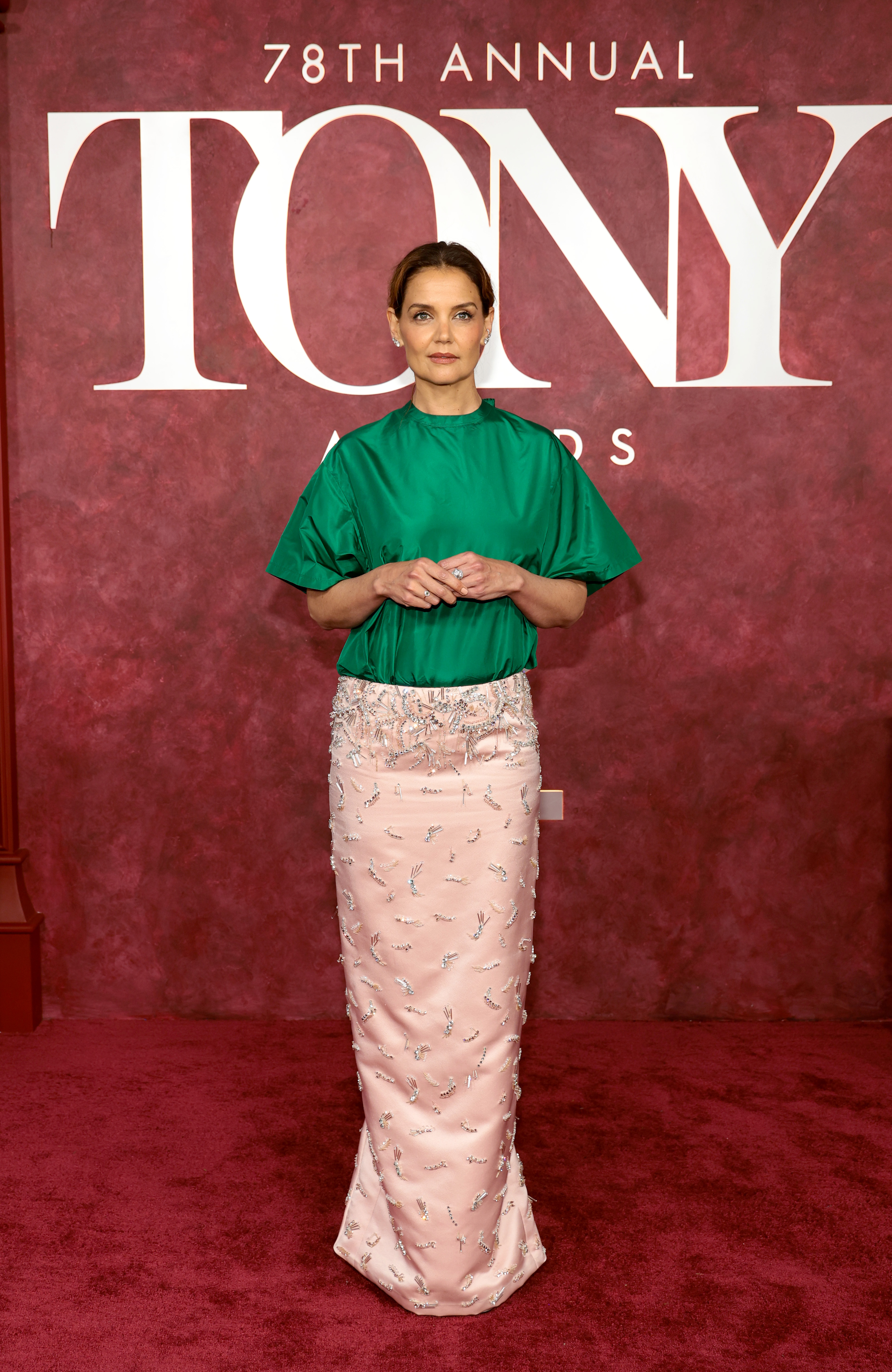 Framed by the official Tony Awards lettering, Katie Holmes takes a wide stance with hands gently clasped. The head-to-toe composition highlights the garment’s unconventional balance of volume, sheen, and ornate detail.