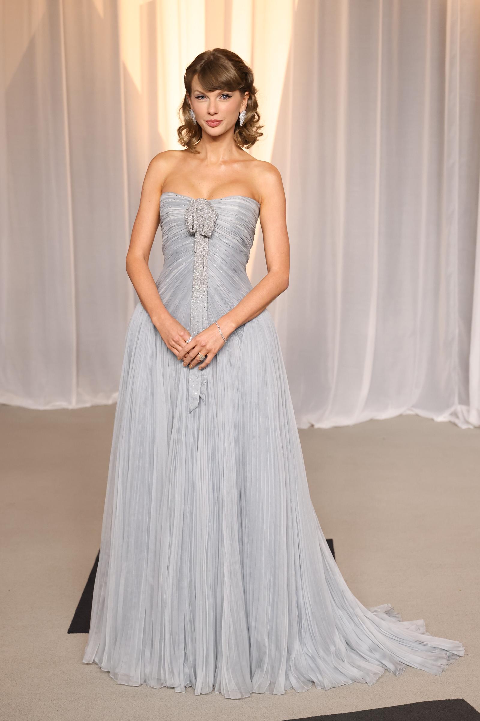 Taylor Swift poses in a strapless, icy-blue pleated gown featuring a crystal-covered bow and sweeping train, accessorized with sparkling earrings, a bracelet, and rings.