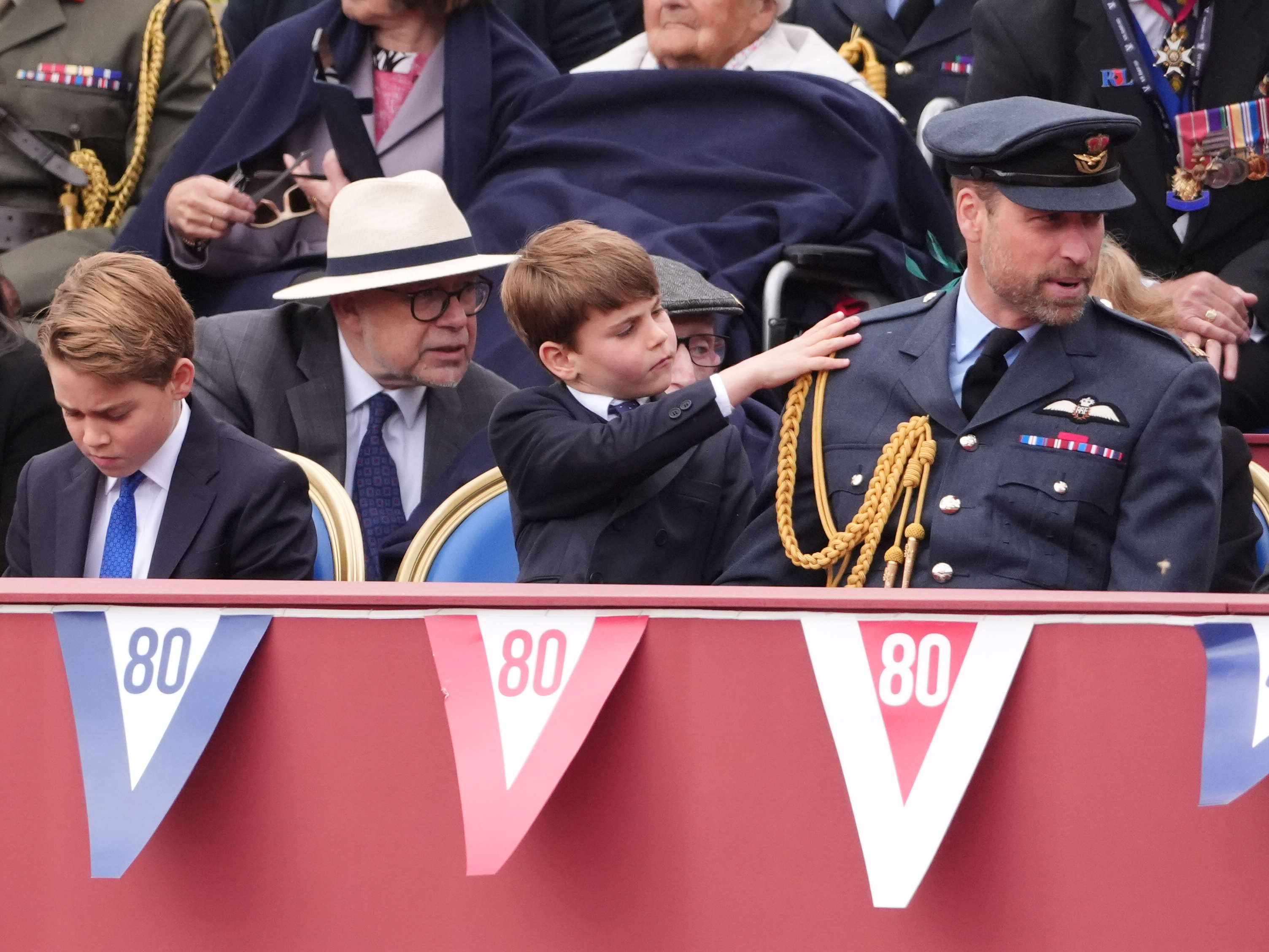 Prince Louis gently brushing off Prince William's RAF No.1 uniform as Prince George sits on the other side of him during the 80th anniversary of VE Day in London, England on May 5, 2025. | Source: Getty Images