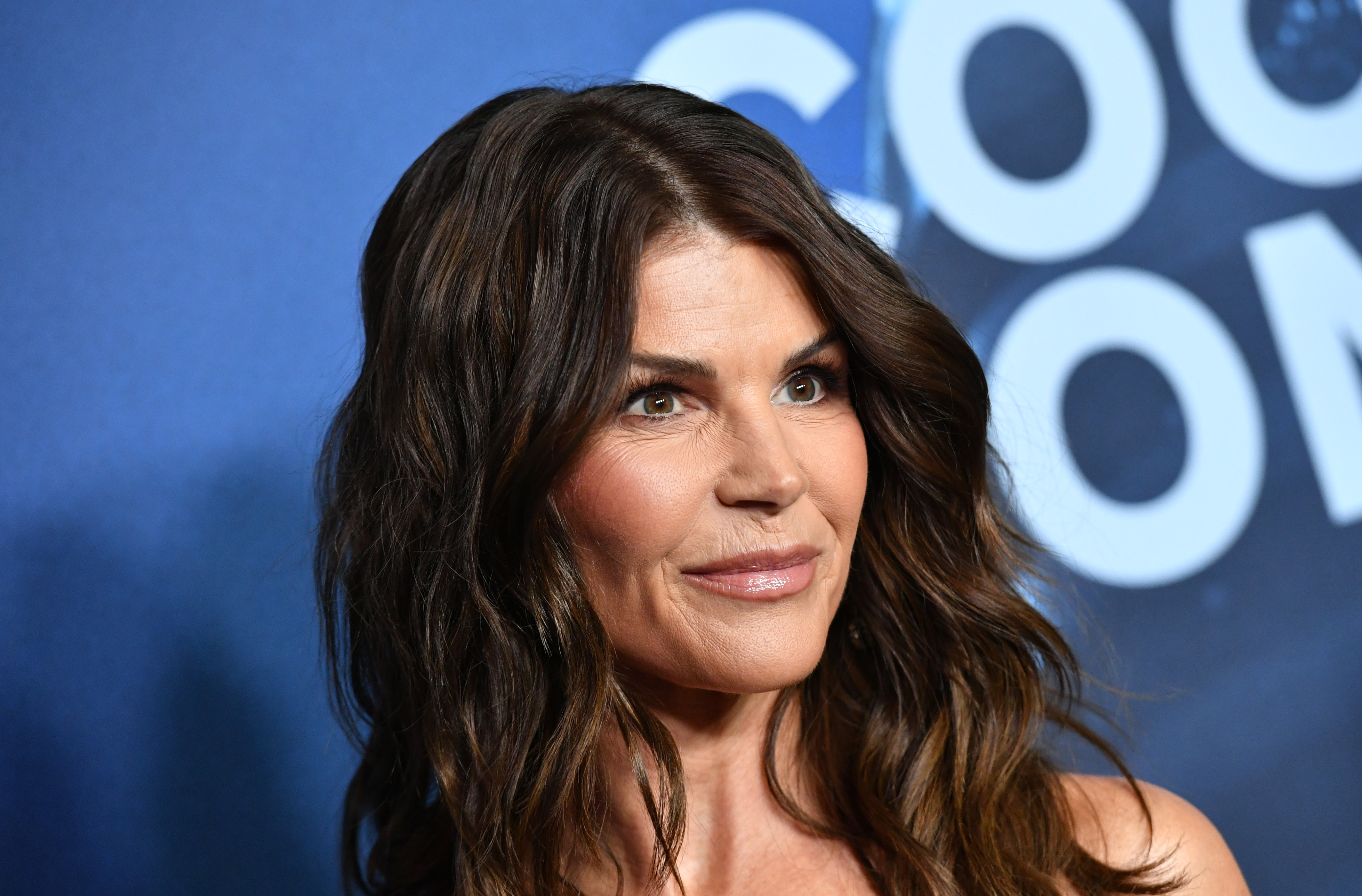 Lori Loughlin smiles radiantly, wearing her brunette hair in soft waves paired with a navy gown accented by gold embroidery. Beloved by television audiences for bringing warmth, grace, and steady wisdom to Aunt Becky on "Full House," the actress highlights enduring poise as she reunites with longtime co-stars to support a cause close to their shared history.
