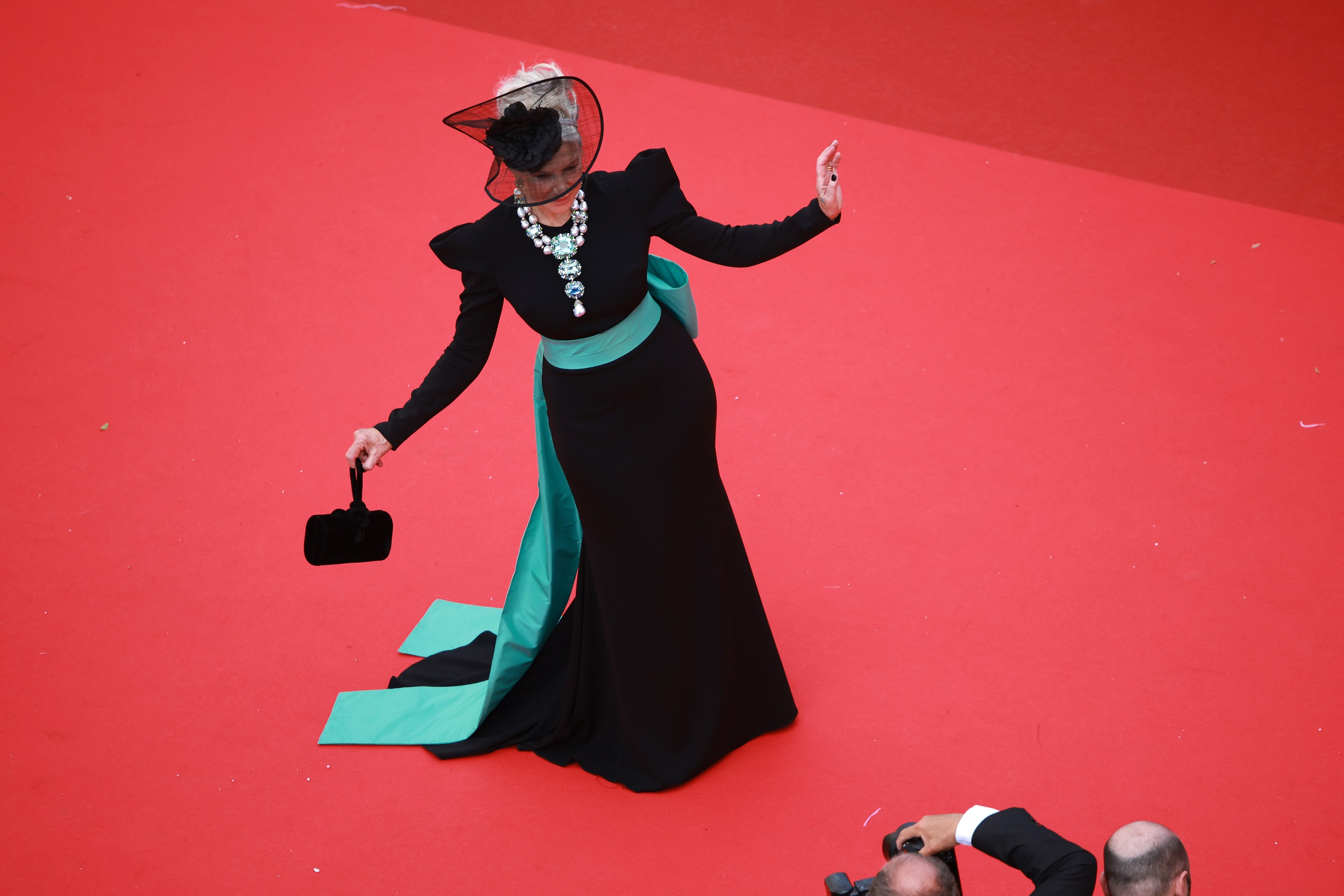 Mirren poses with theatrical flair, one arm outstretched, the other holding a bag. Her aquamarine sash and sparkling necklace pop vividly against the red carpet, turning her into a living jewel amid the bold contrast.