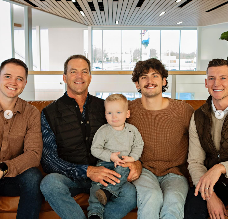 Bryon Noem sits on a couch with three younger men and a toddler, all smiling toward the camera in a bright, modern indoor space with large windows behind them. | Source: Instagram/kristinoem