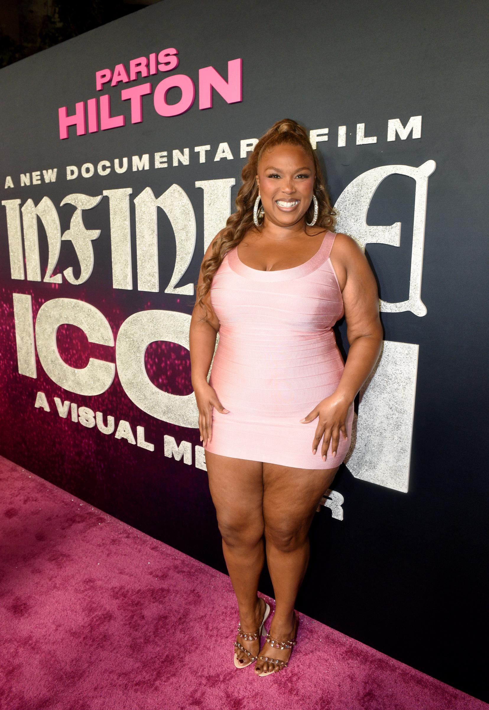 Standing in front of the event signage, she grins broadly with both hands placed at her waist. Her midsection appears firm and cinched, with minimal bloating or curvature at the belly, highlighting her weight loss transformation.