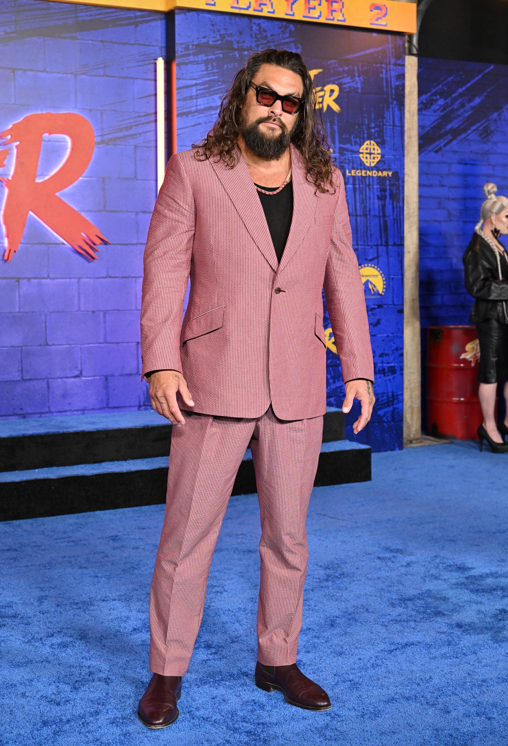 Standing full-length on the vibrant blue carpet, Jason Momoa completes the look with straight-leg trousers tailored to break neatly over polished deep burgundy Chelsea boots. A metal barrel prop and event branding frame the arcade-inspired backdrop behind him as he faces the photographers head-on. | Source: Getty Images