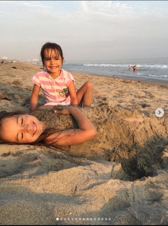 Mabel and Fiona are pictured as young sisters on a sunny beach, with Fiona playfully buried in the sand as Mabel smiles behind her. The carefree childhood moment now carries a poignant weight following Fiona’s death. | Source: Instagram/mabellowe_