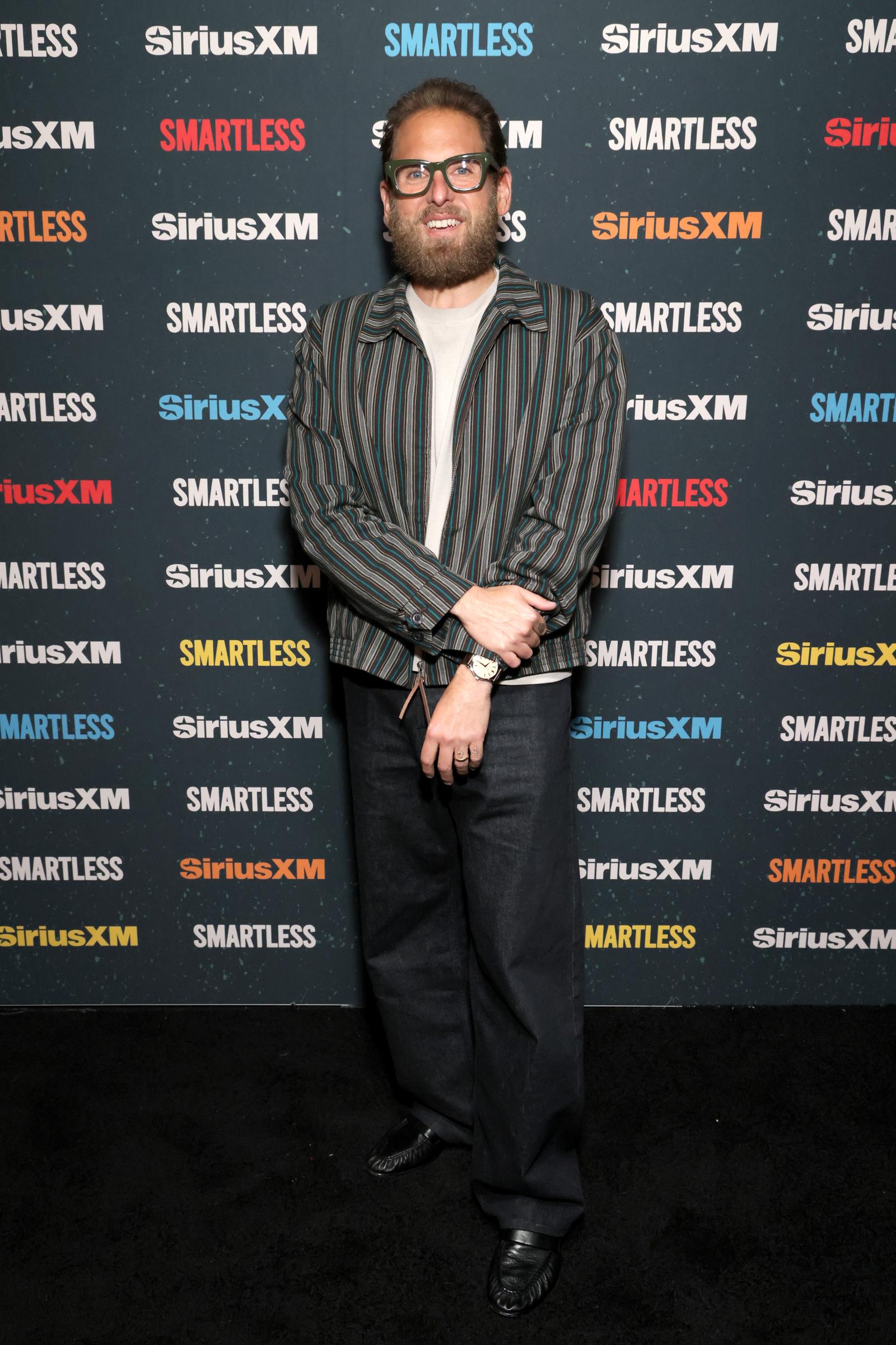 Jonah Hill attended SiriusXM's "SmartLess LIVE" at Avalon Hollywood in Los Angeles on April 25, 2026, where he posed against the show's colorful step-and-repeat backdrop. Wearing a striped jacket over a light shirt with loose dark trousers, black shoes, and bold glasses, Hill sported a full beard and longer hair while displaying his notably slim frame. The appearance offered one of the latest looks at his transformation, years after his changing weight.