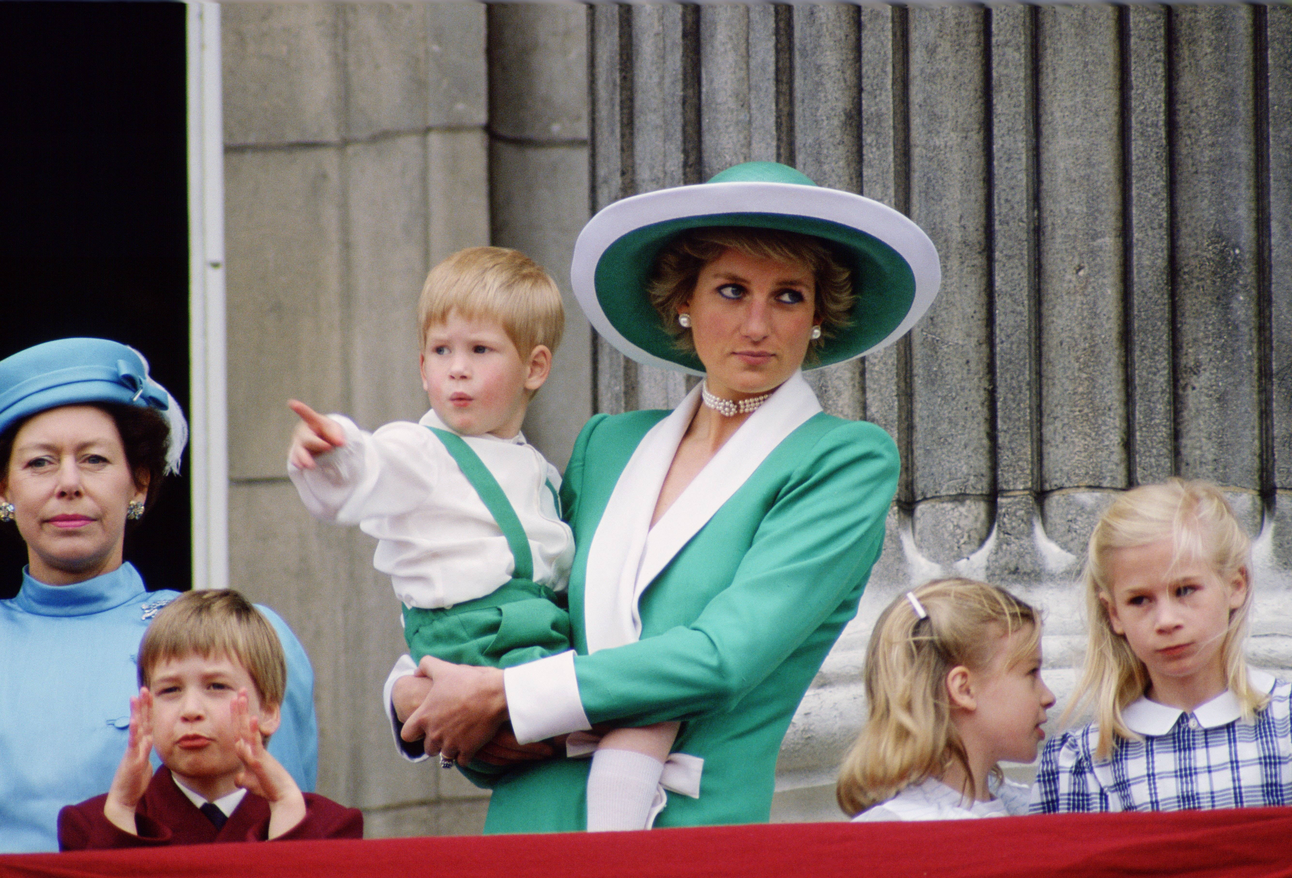 Princess Diana, dressed in a green and white suit with a matching hat, holds a young Prince Harry in her arms as she watches the 1988 Trooping the Colour from the Buckingham Palace balcony. Surrounded by Prince William, Lady Rose Windsor, Lady Davina Windsor, and Princess Margaret, Diana’s composed presence captures a timeless royal family moment framed by tradition and pageantry.
