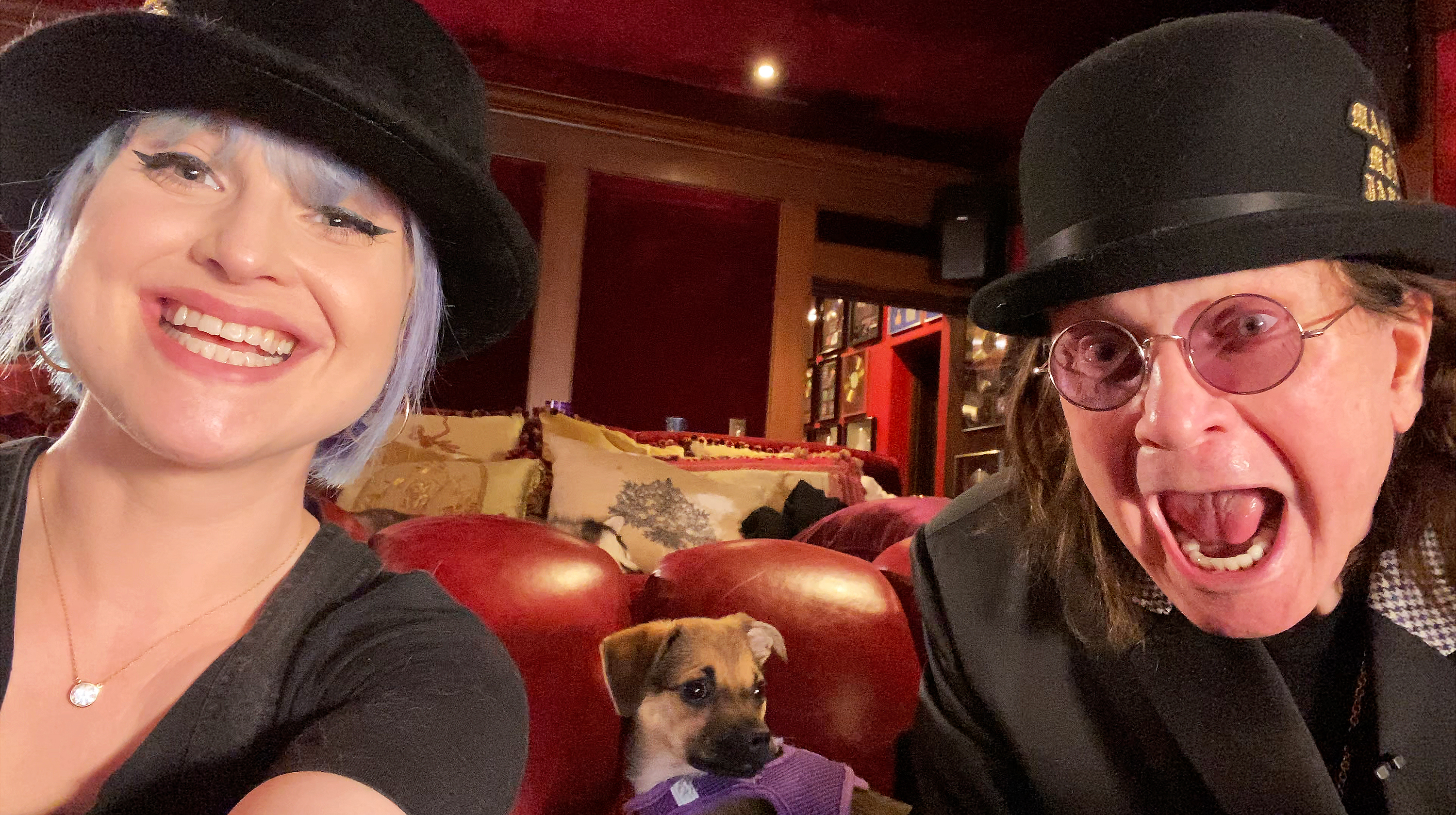 Kelly and Ozzy smile for a selfie while filming "Celebrity Watch Party." Kelly wears a black hat and light blue hair, while Ozzy opens his mouth in a playful expression. A small dog sits between them on the couch. | Source: Getty Images
