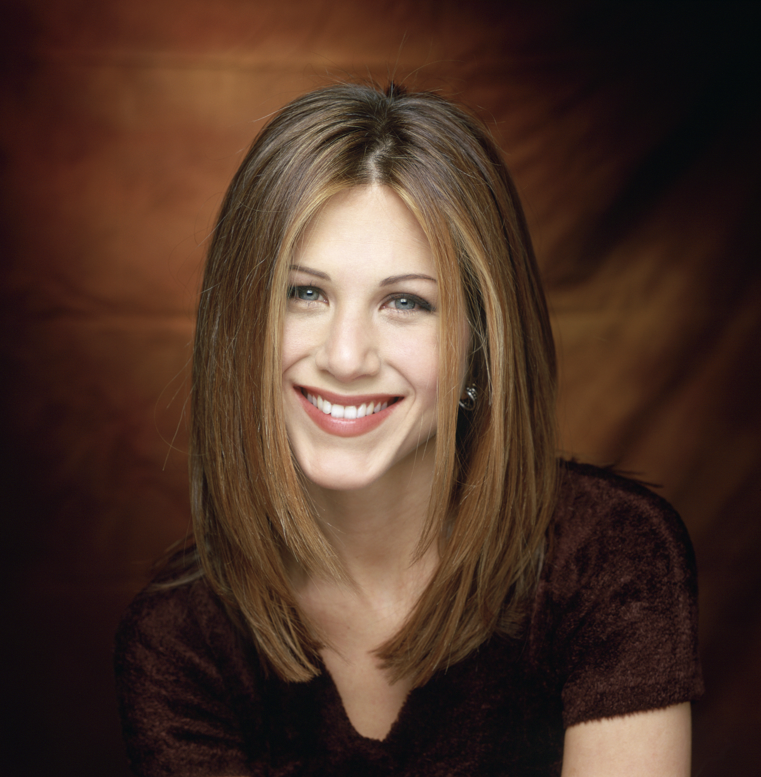 Jennifer Aniston as Rachel Green, smiles warmly with her iconic straight, layered hairstyle. She wears a dark top against a softly lit brown background, radiating charm and confidence.