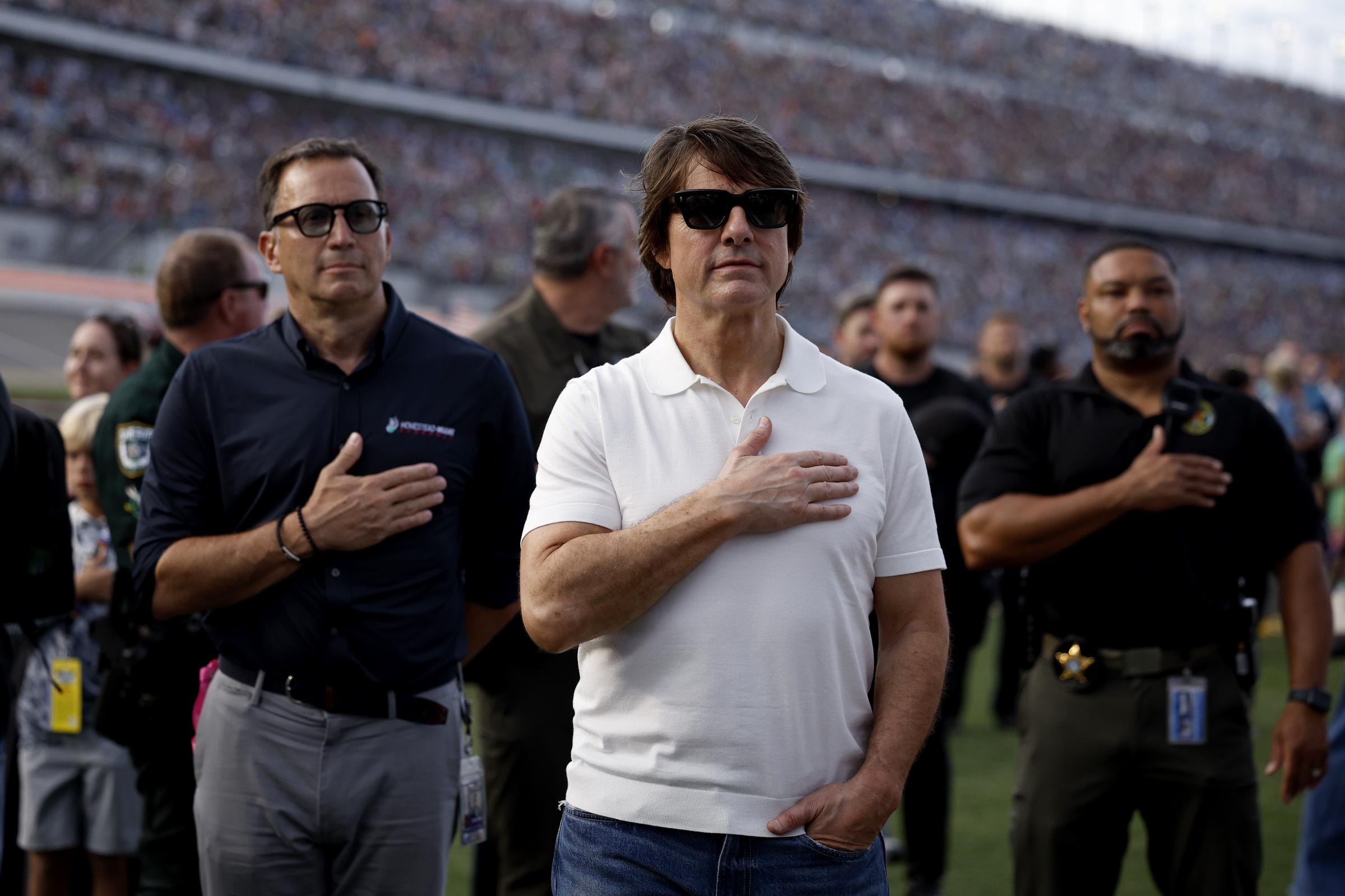 Cruise stands at Daytona International Speedway with his right hand over his chest as others around him do the same. He wears a white polo shirt, blue jeans and dark sunglasses, with a packed grandstand visible behind the group. | Source: Getty Images