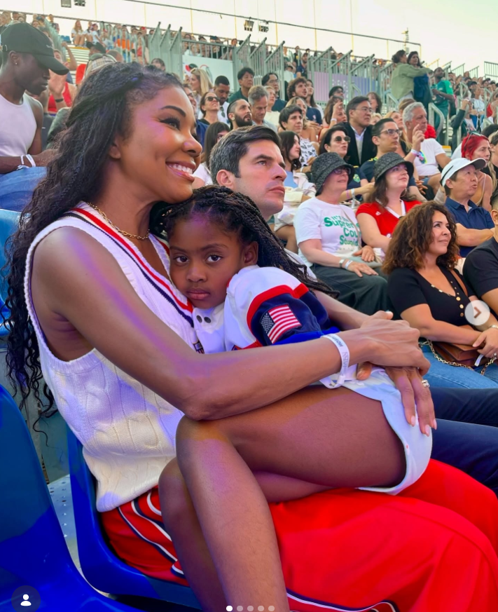 Gabrielle Union sits in stadium seating holding her daughter, Kaavia James Union Wade, on her lap. Gabrielle smiles warmly while wearing a sporty white sleeveless top and red pants, as Kaavia rests against her with a calm, slightly serious expression. | Source: Instagram/gabunion