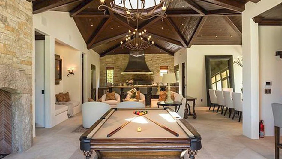The poolside entertainment pavilion pairs rustic stone walls and a cozy fireplace with a billiards table, dramatic vaulted wood ceilings, a built-in outdoor kitchen with a metal vent hood, and an expansive dining table setup. | Source: YouTube/Taste of Country