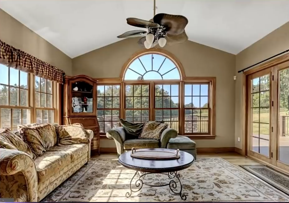 Large windows with warm wooden frames and an arched centerpiece dominate this living room, bringing natural light into the space. A floral-patterned sofa, a matching armchair, a decorative rug, and a wooden display cabinet complete the traditionally styled interior.