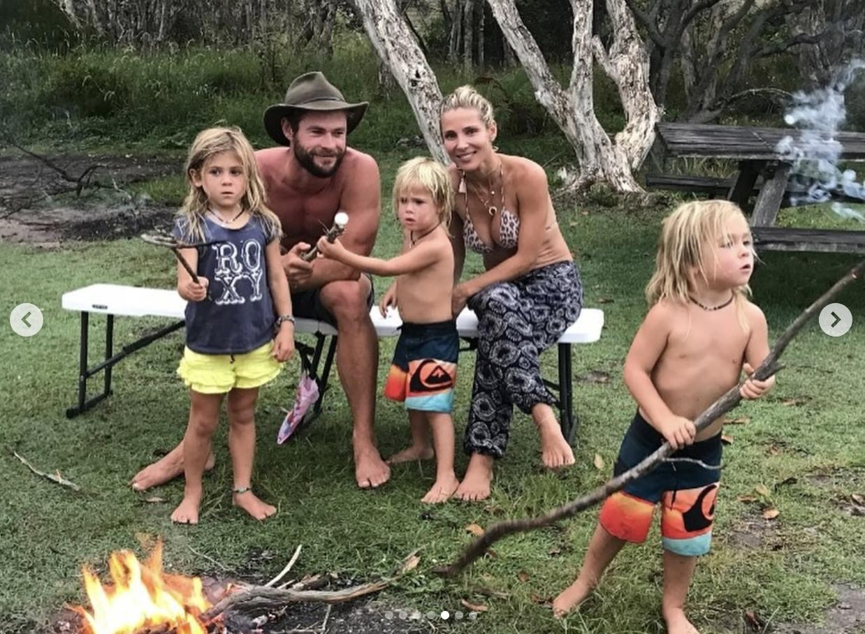 Chris Hemsworth and Elsa Pataky enjoy a relaxed outdoor moment with their three children — twins Tristan and Sasha, and daughter India Rose. The family is pictured barefoot and gathered around a small campfire, roasting marshmallows and holding sticks, with a forested backdrop behind them.