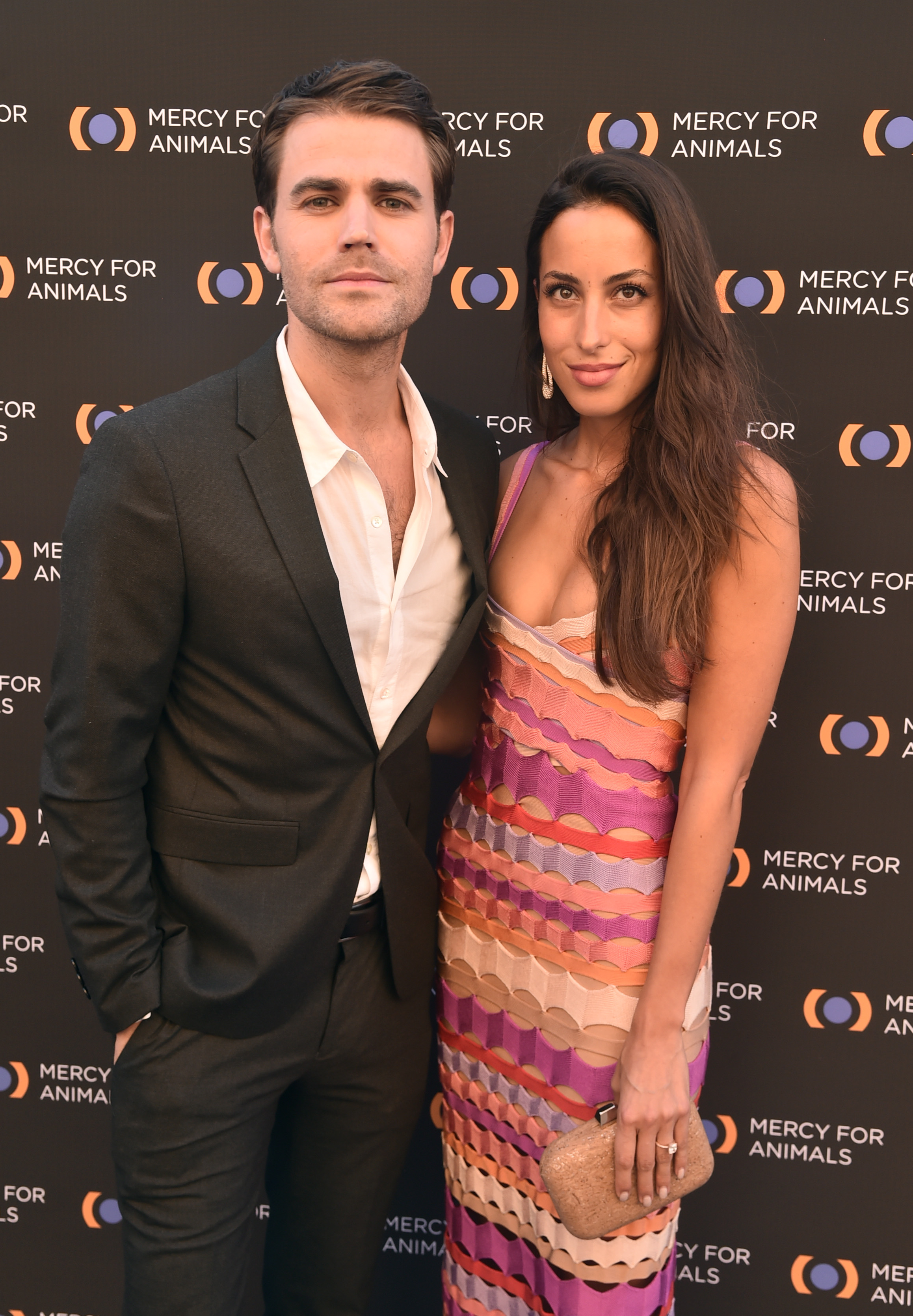 Paul Wesley and Ines de Ramon attended the Mercy For Animals 20th Anniversary Gala at The Shrine Auditorium on September 14, 2019, in Los Angeles, California. Wesley looked dapper in a tailored black suit paired with an open-collar white dress shirt, posing alongside de Ramon, who wore a colorful chevron-striped crochet slip dress in shades of purple, orange, and cream, accessorized with a sparkling clutch and delicate earrings against the event step-and-repeat. | Source: Getty Images
