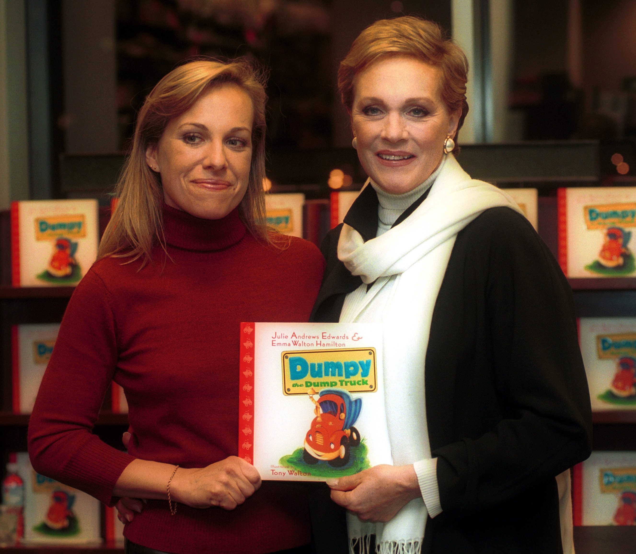 Julie Andrews and her daughter, Emma Walton Hamilton, proudly display their collaborative children's book "Dumpy the Dump Truck" during a signing event at Barnes & Noble in New York City on November 28, 2000. Andrews smiles warmly in a black jacket layered over an ivory turtleneck with a white scarf and gold hoop earrings, holding the illustrated book alongside Hamilton, who wears a burgundy turtleneck sweater.