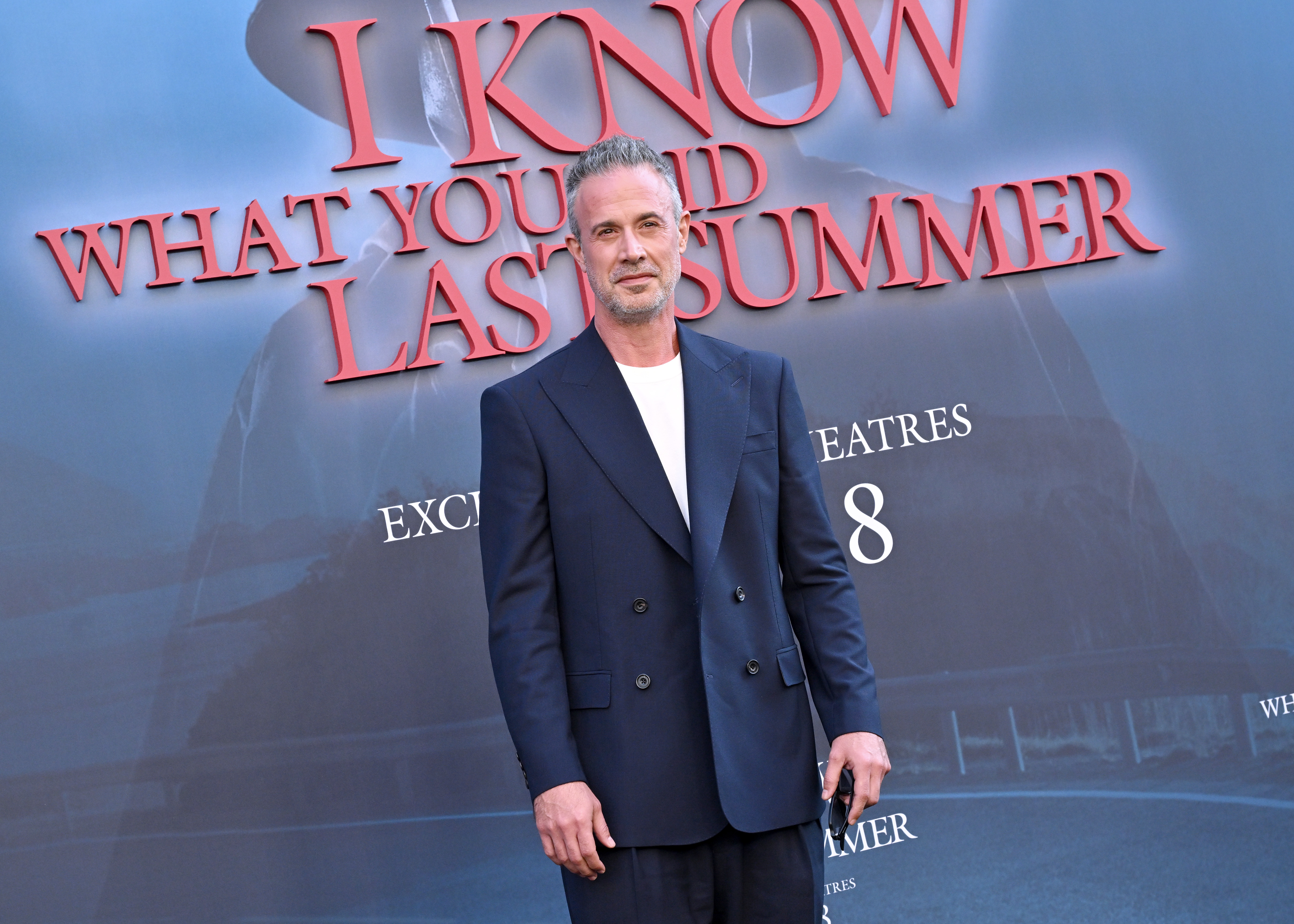 Prinze Jr.'s outfit is more visible here as well with his navy blazer over a white shirt and matching pants. His expression is relaxed and self-assured as he poses in front of the red carpet poster for "I Know What You Did Last Summer," which features bold red lettering over a dark, moody landscape with a shadowy figure in the background.