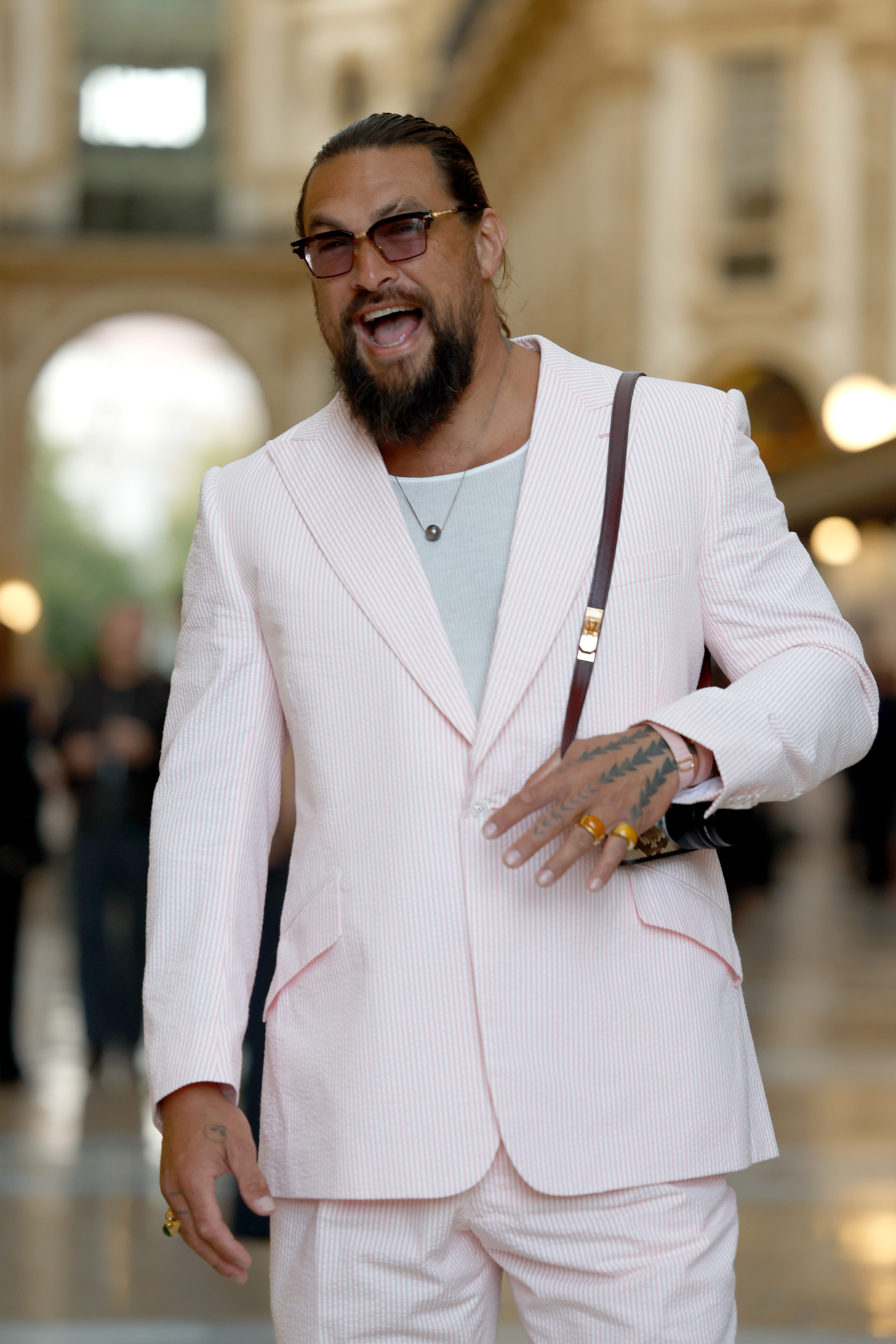 Momoa laughs while posing inside the venue, offering a closer look at his tinted glasses, jewelry and the fine pink stripes running through his suit. | Source: Getty Images