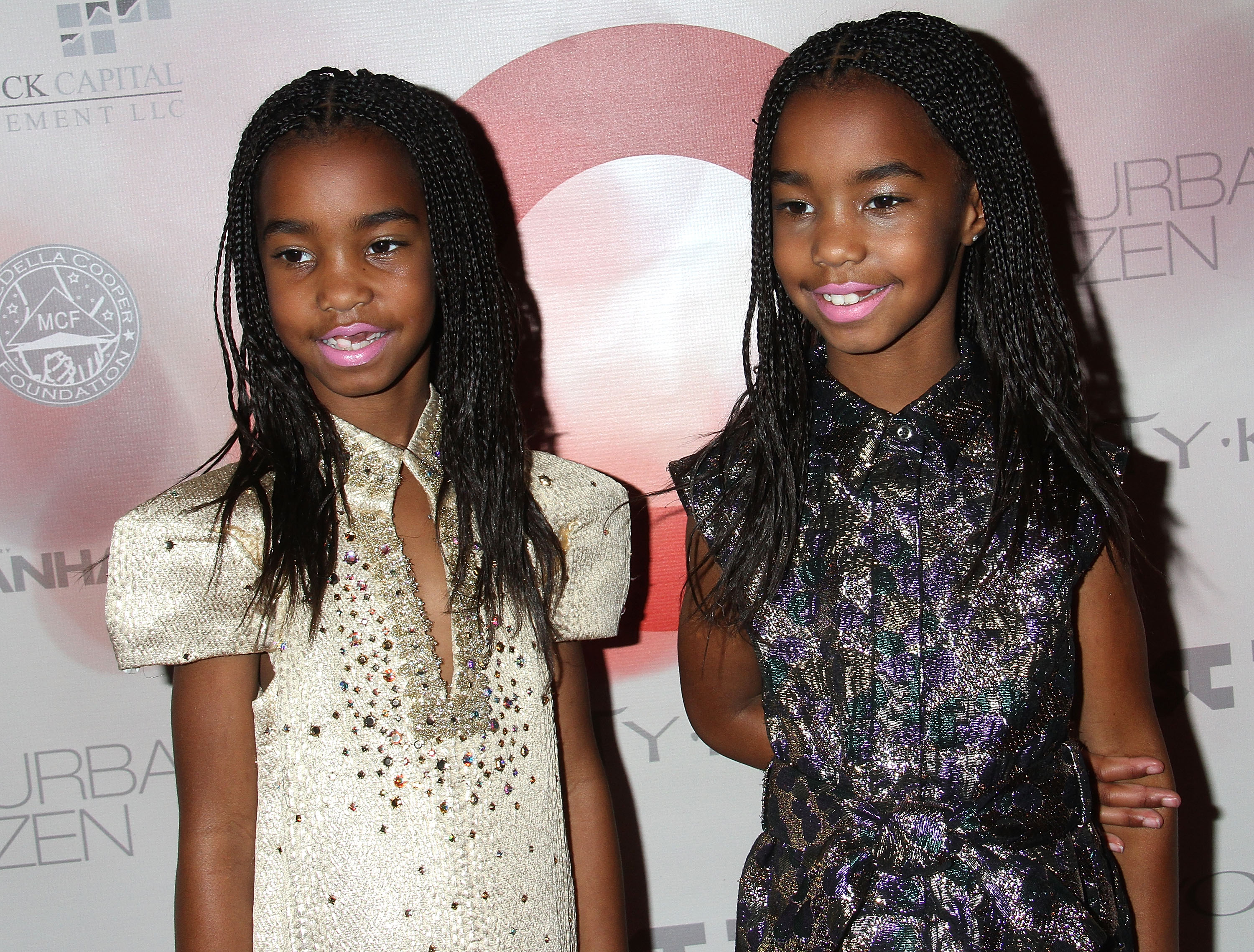 A young Jessie and D'Lila Combs pose side by side in sparkling, fashion-forward outfits, their braided hairstyles and bright lip colors adding a playful polish. Their confident smiles and coordinated glamour stand out against the event backdrop, giving the moment a radiant charm.