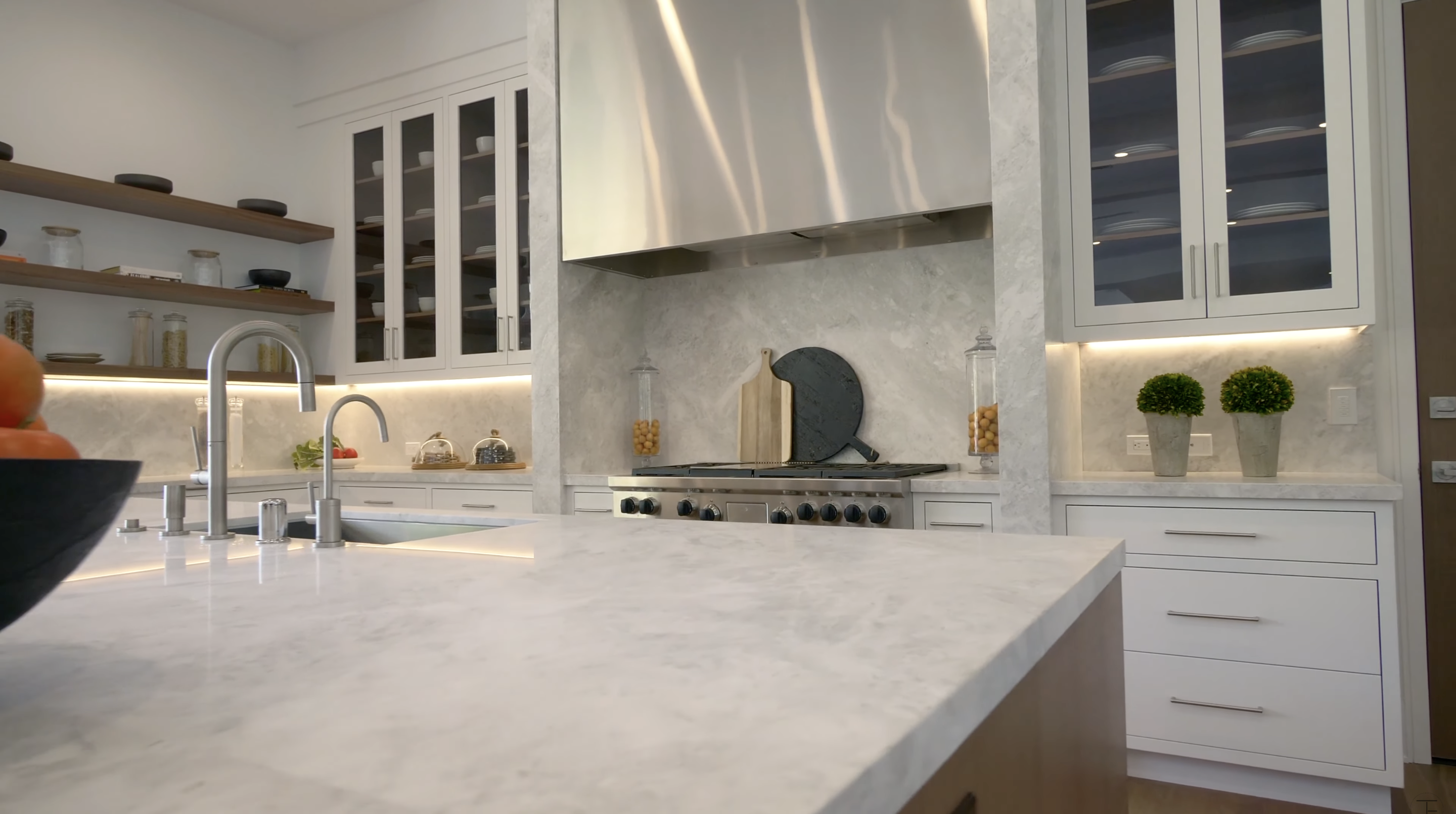 White cabinetry and pale stone dominate the kitchen, while open wood shelving adds a warmer finish. A large island fills the foreground, with a stainless-steel range and hood defining the cooking area. | Source: Youtube/Tomer.Fridman