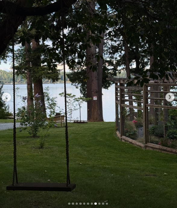 A simple wooden swing hangs suspended from a tree branch overlooking the tranquil, tree-lined shores of Vancouver Island at Arcady. In the background, tall coastal conifers frame the glistening water beside enclosed garden fencing designed to protect Pamela's growing beds, capturing the unhurried serenity of her seaside retreat. She accompanied the image with the caption, "I think of life as a garden… You get to replant, start again, and make it yours. 🤍 Love, P". | Source: Instagram/pamelaanderson