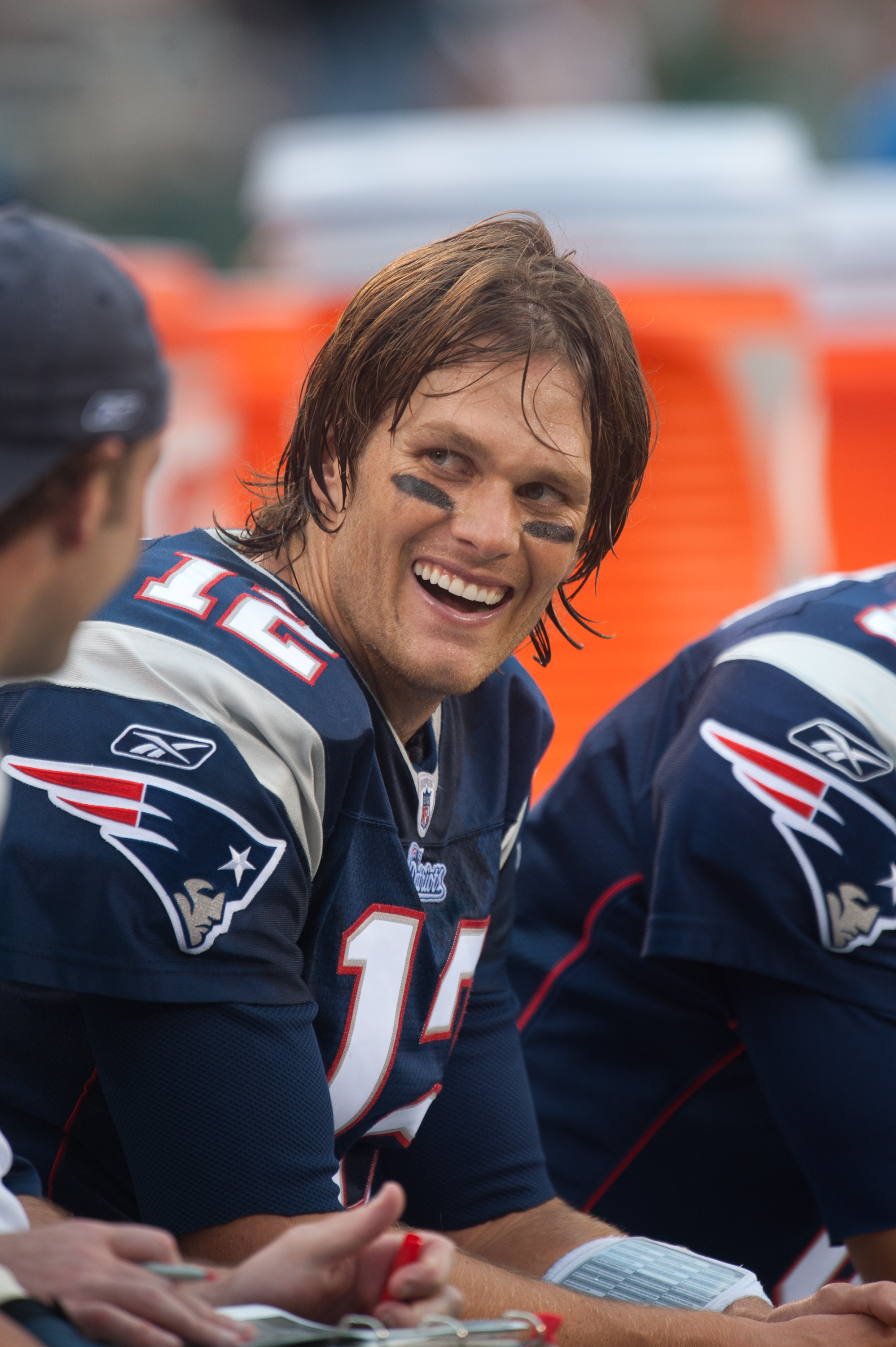 Teeth flashing in a bright, confident smile, Tom Brady looks on from the bench during a matchup against the New York Jets at New Meadowlands Stadium in East Rutherford, New Jersey. He is happy and clearly in his element.