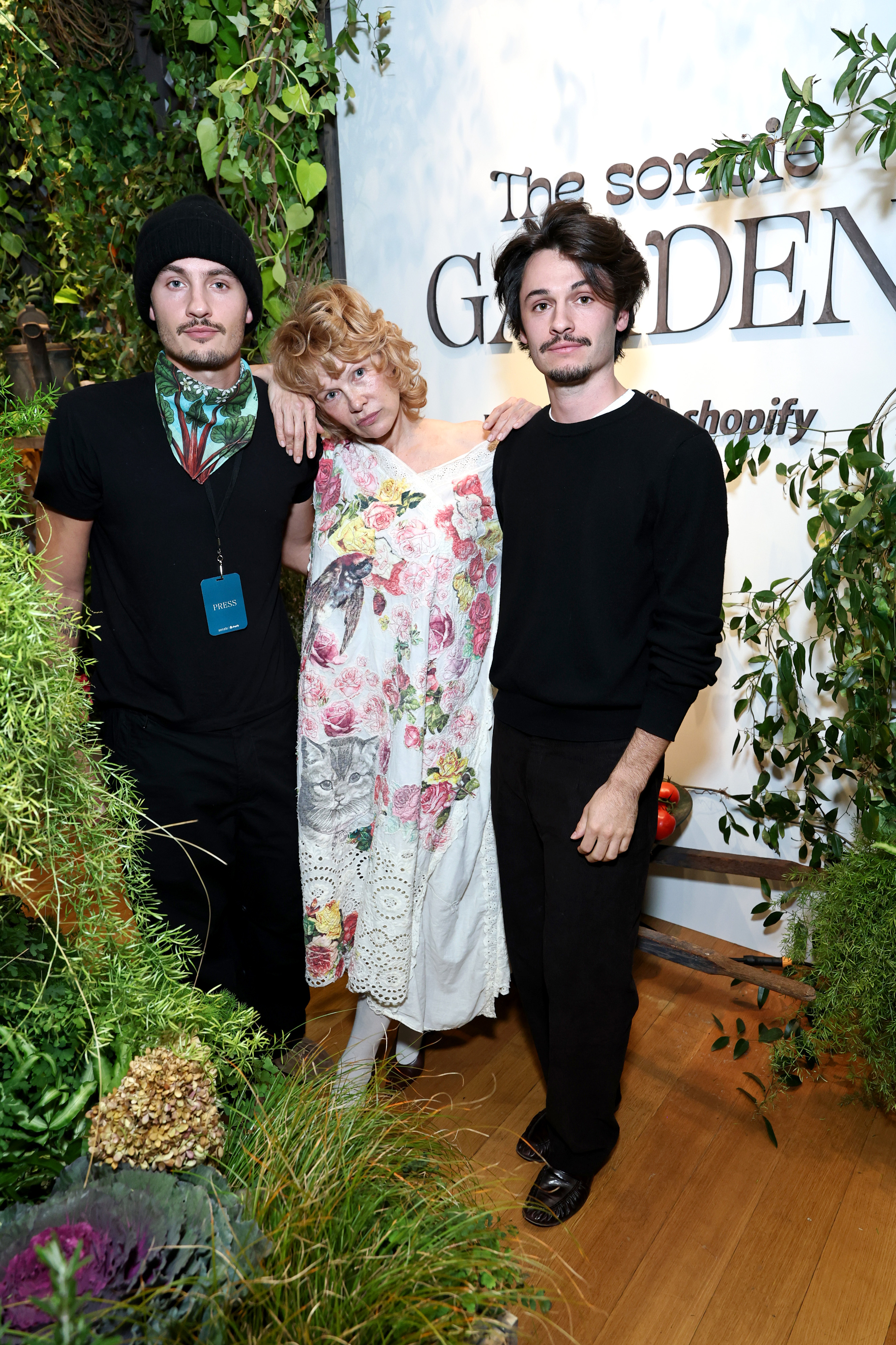 The trio at the Sonsie Garden event in New York City in 2025. Wearing a white floral dress detailed with birds, cats, and roses, Anderson stands with her arms gently draped around her sons. Dylan wears a black beanie and printed scarf, while Brandon opts for a simple black crewneck. Surrounded by greenery and natural textures, the trio presents a striking image of family presence and quiet unity.
