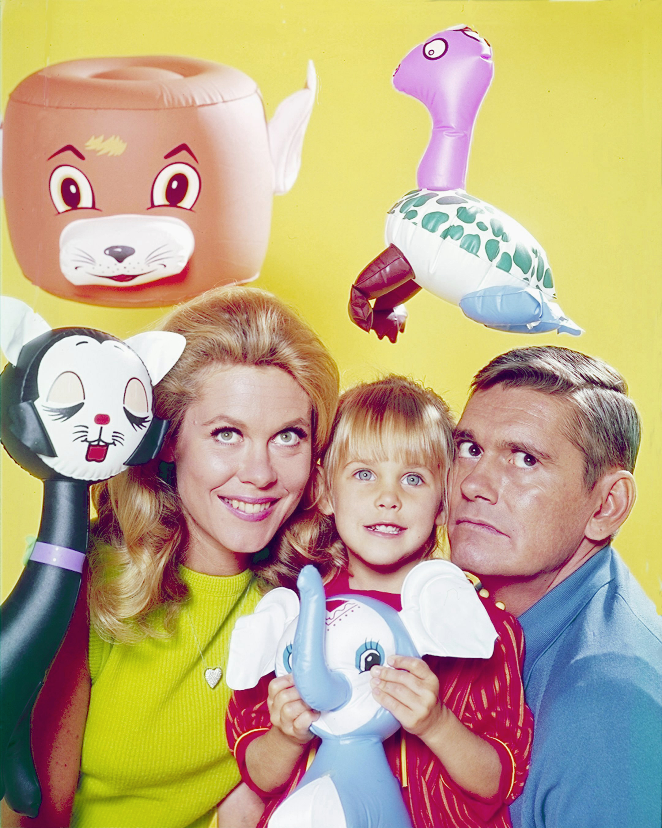 Erin Murphy poses between Elizabeth Montgomery and Dick York in a colorful "Bewitched" promotional image surrounded by oversized toy figures. Murphy is still a small child here, making the photo a sharp contrast with the polished selfies fans are seeing from her six decades later.